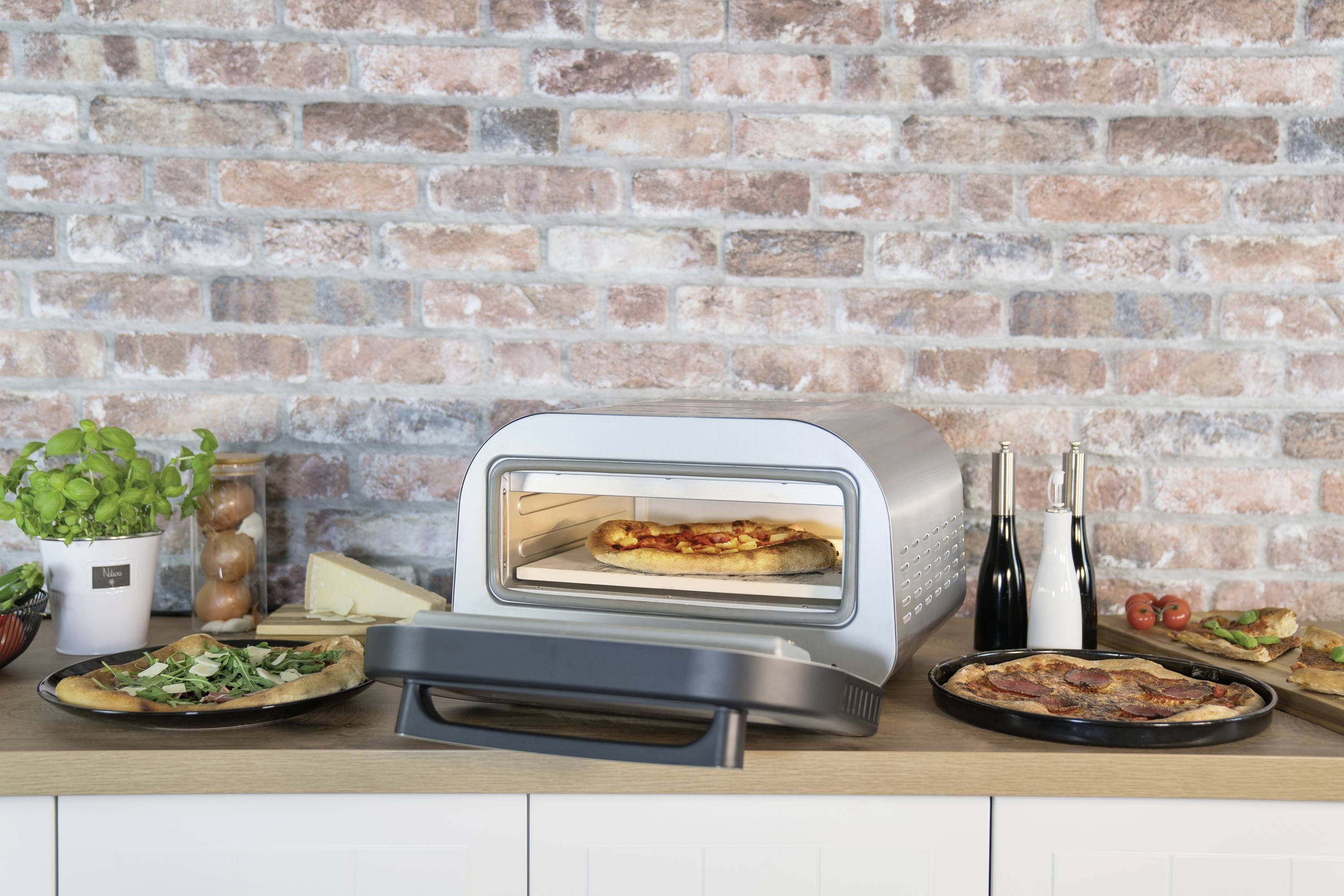 Un forno per pizza su un piano da lavoro cucina sta cuocendo una pizza. Accanto ci sono altre pizze sui piatti. Sullo sfondo si trova un muro di mattoni.