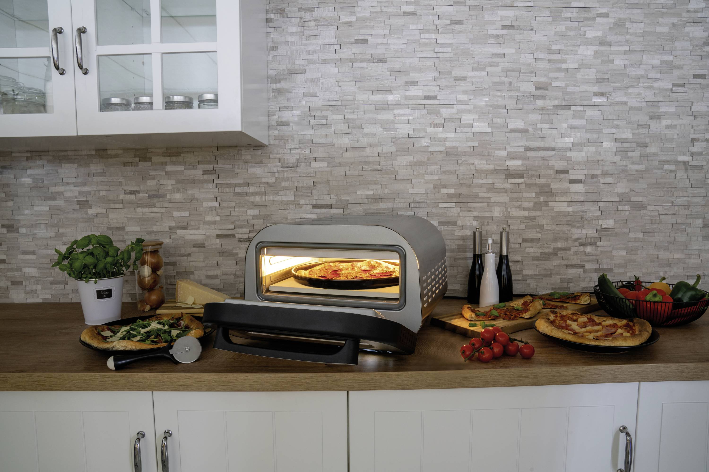 Un forno per pizza su un bancone della cucina sta cuocendo una pizza. Accanto ci sono ingredienti freschi come basilico, pomodori e altre verdure.