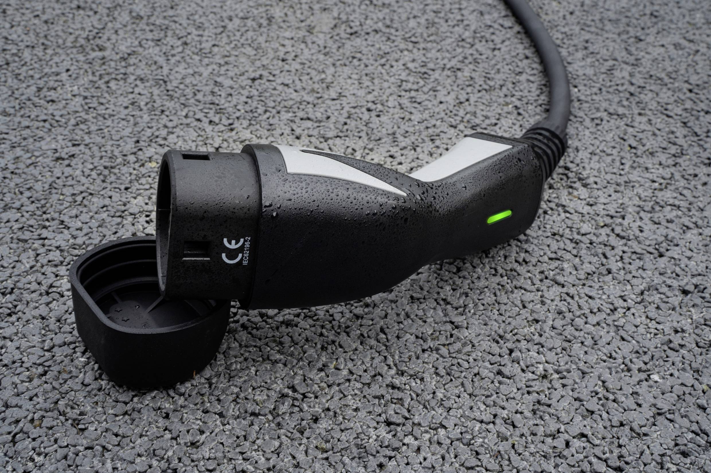 Primo piano di un connettore di ricarica per veicolo elettrico su asfalto bagnato. Una luce verde è accesa sul connettore.