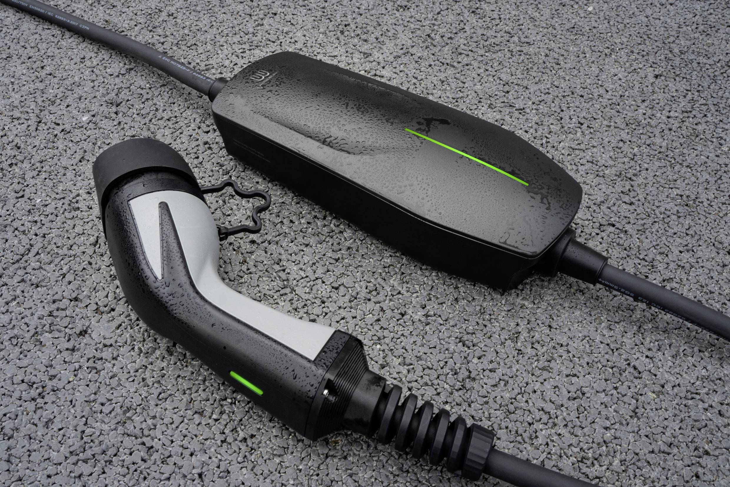 Un cavo di ricarica per auto elettrica è posizionato su una superficie asfaltata. Sono visibili una spina e un caricabatterie con indicatori LED verdi.