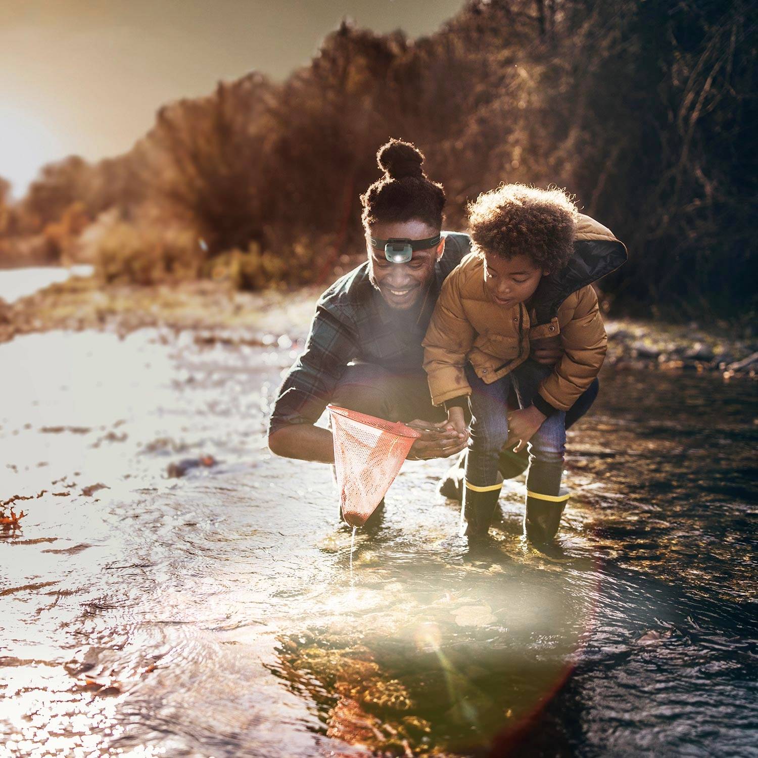 Una persona con un bambino raccoglie acqua con una rete in un fiume al tramonto. Entrambi indossano stivali di gomma.