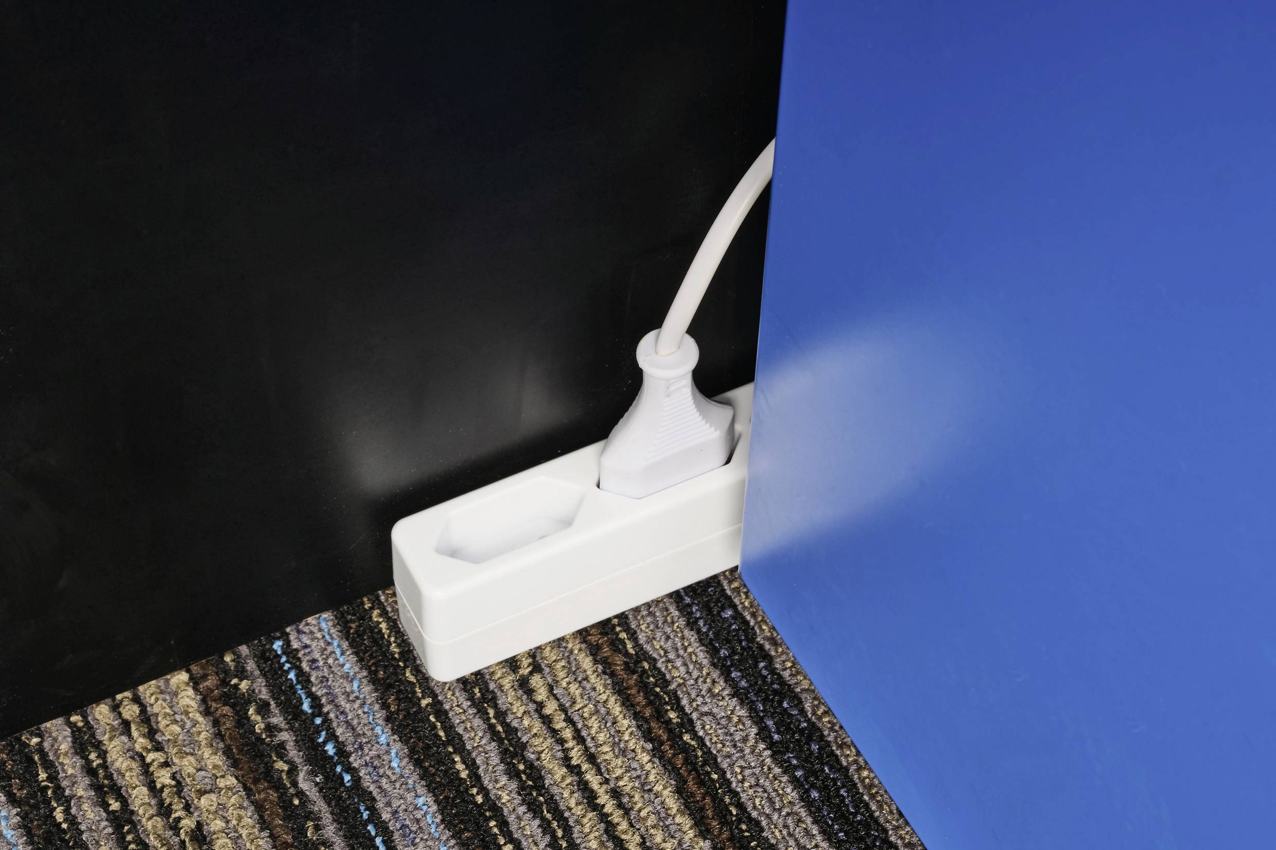 Un cavo di prolunga bianco con una spina inserita è posizionato su un tappeto a righe tra una parete nera e una blu.