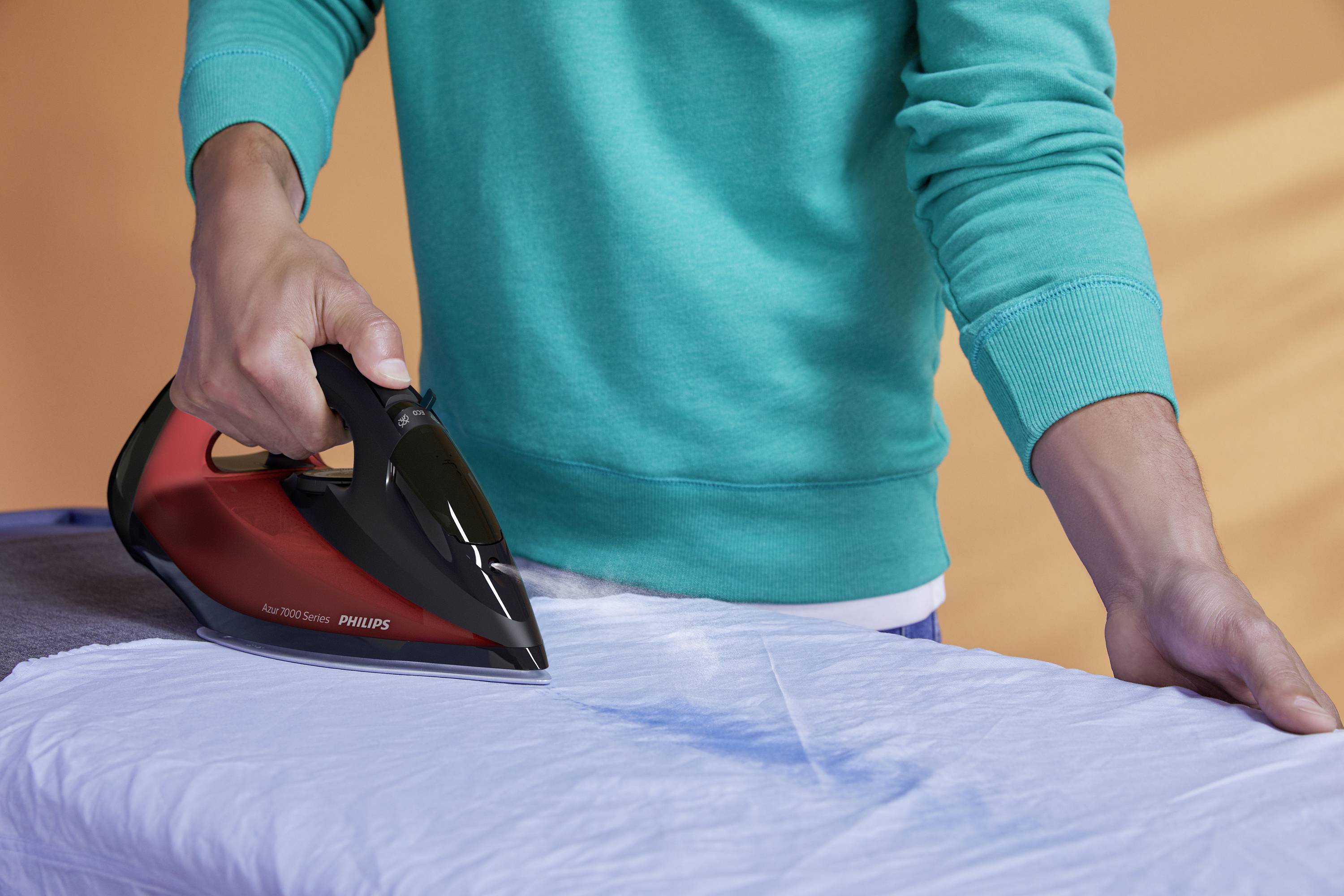 Una persona sta stirando una camicia bianca con un ferro da stiro nero e rosso su un asse da stiro, sullo sfondo toni caldi.