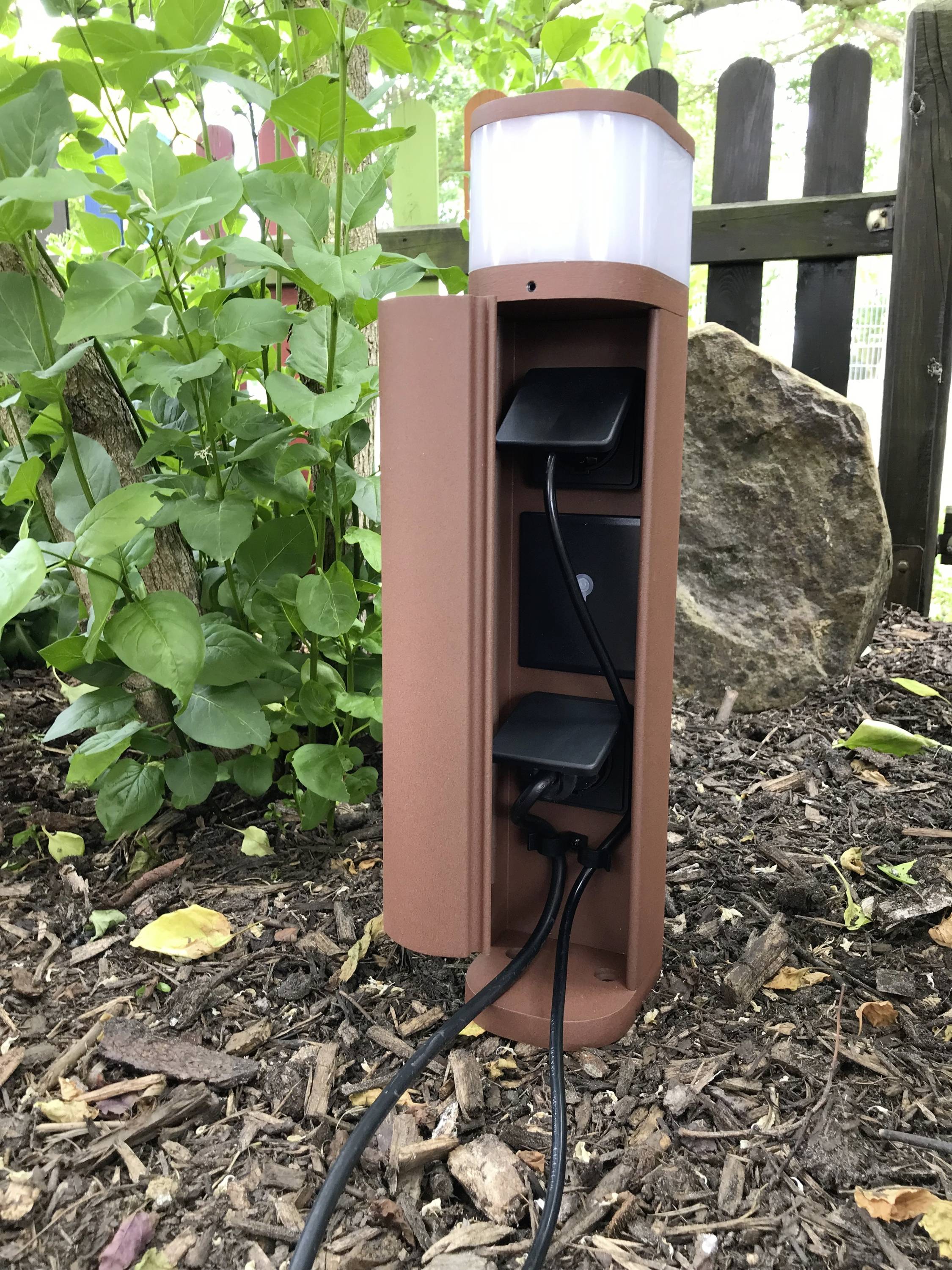 Una presa elettrica marrone è collocata in giardino. Ha due slot con cavi inseriti ed è circondata da piante verdi.