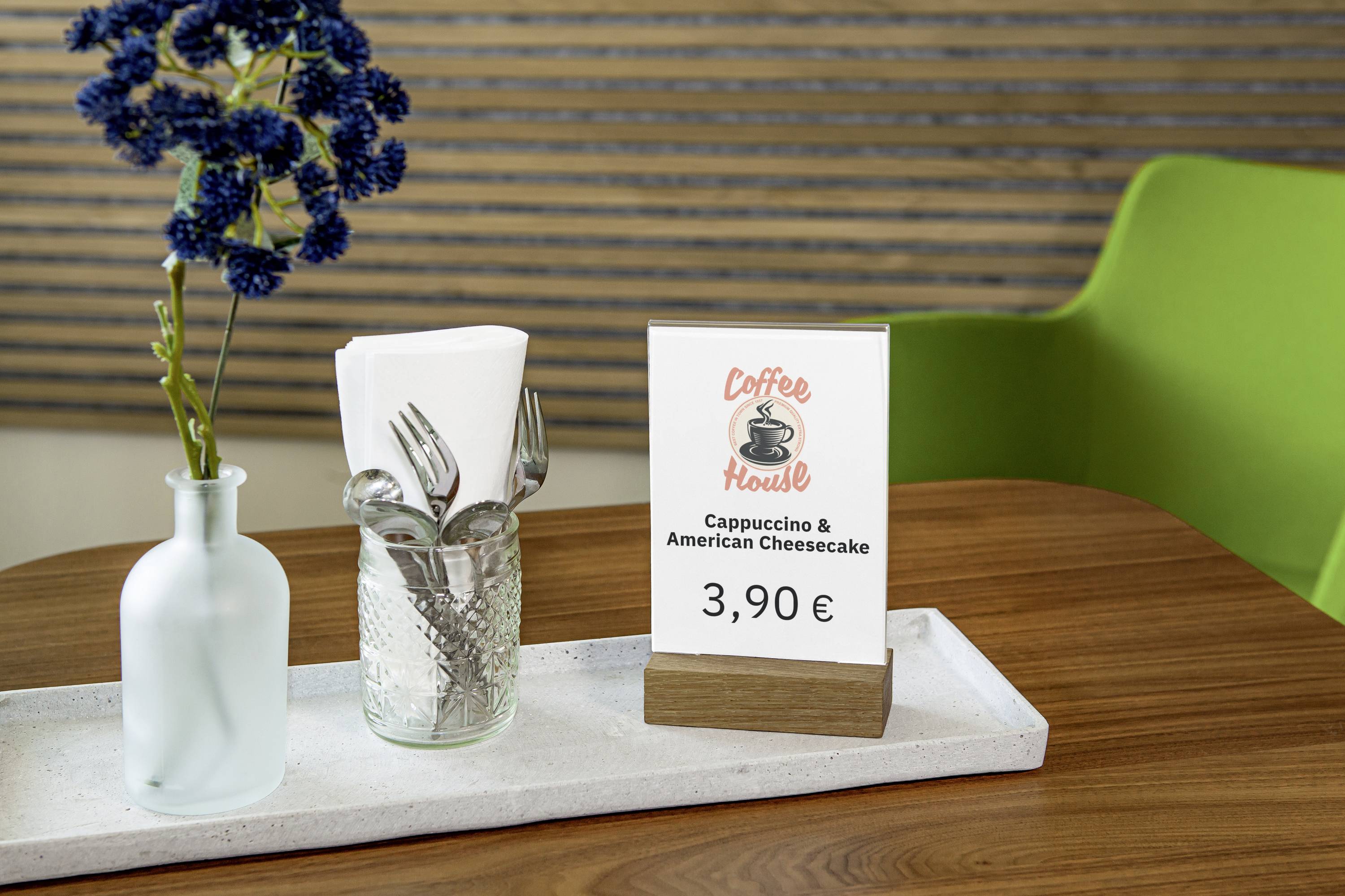 Un tavolo con un bicchiere e delle posate, un vaso con fiori e un espositore per un 'Cappuccino & American Cheesecake' a 3,90 EUR.
