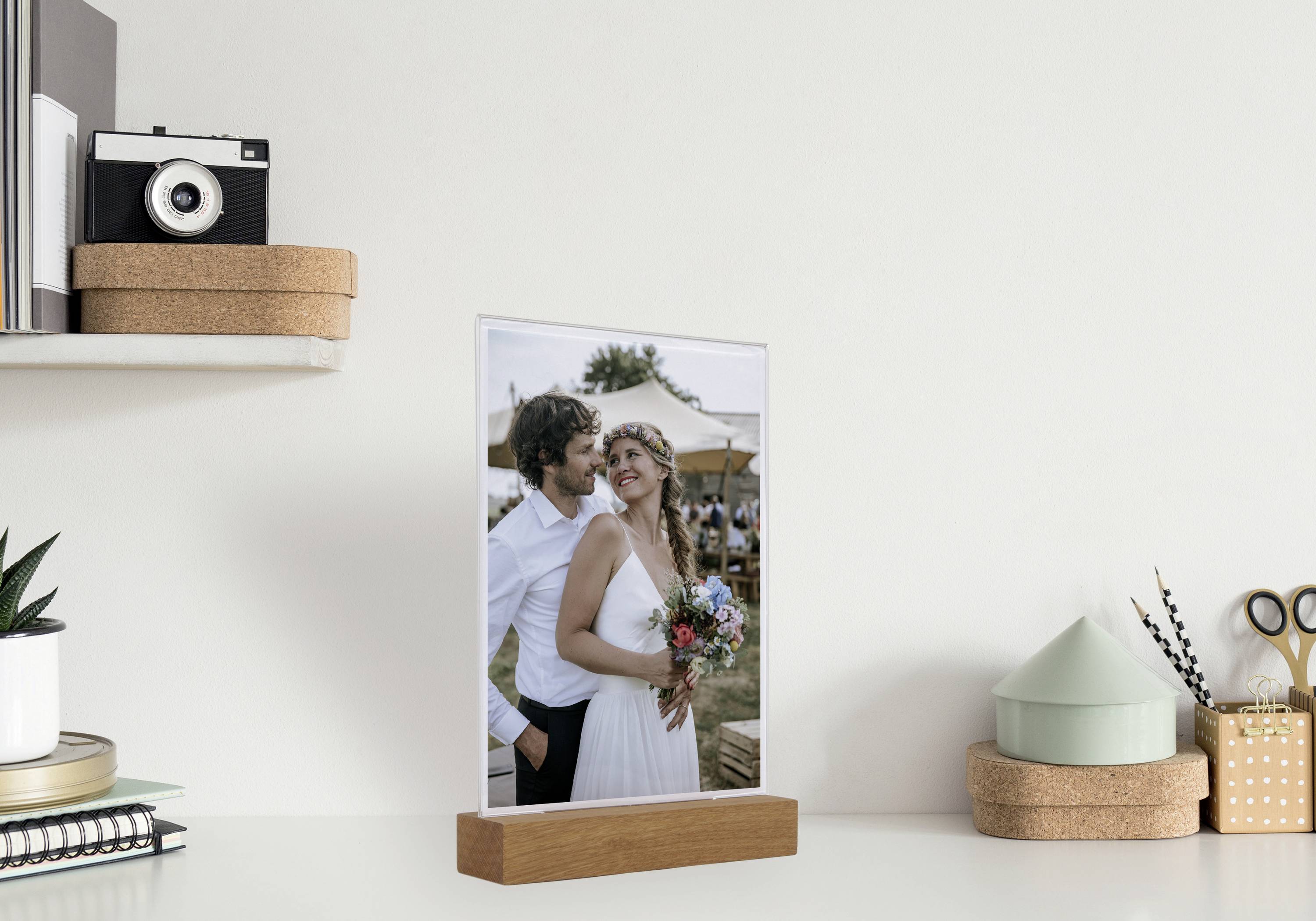 Una foto in una cornice di legno mostra una coppia vestita da sposi. Stanno insieme sorridendo. Sullo sfondo si intravvedono festeggiamenti.