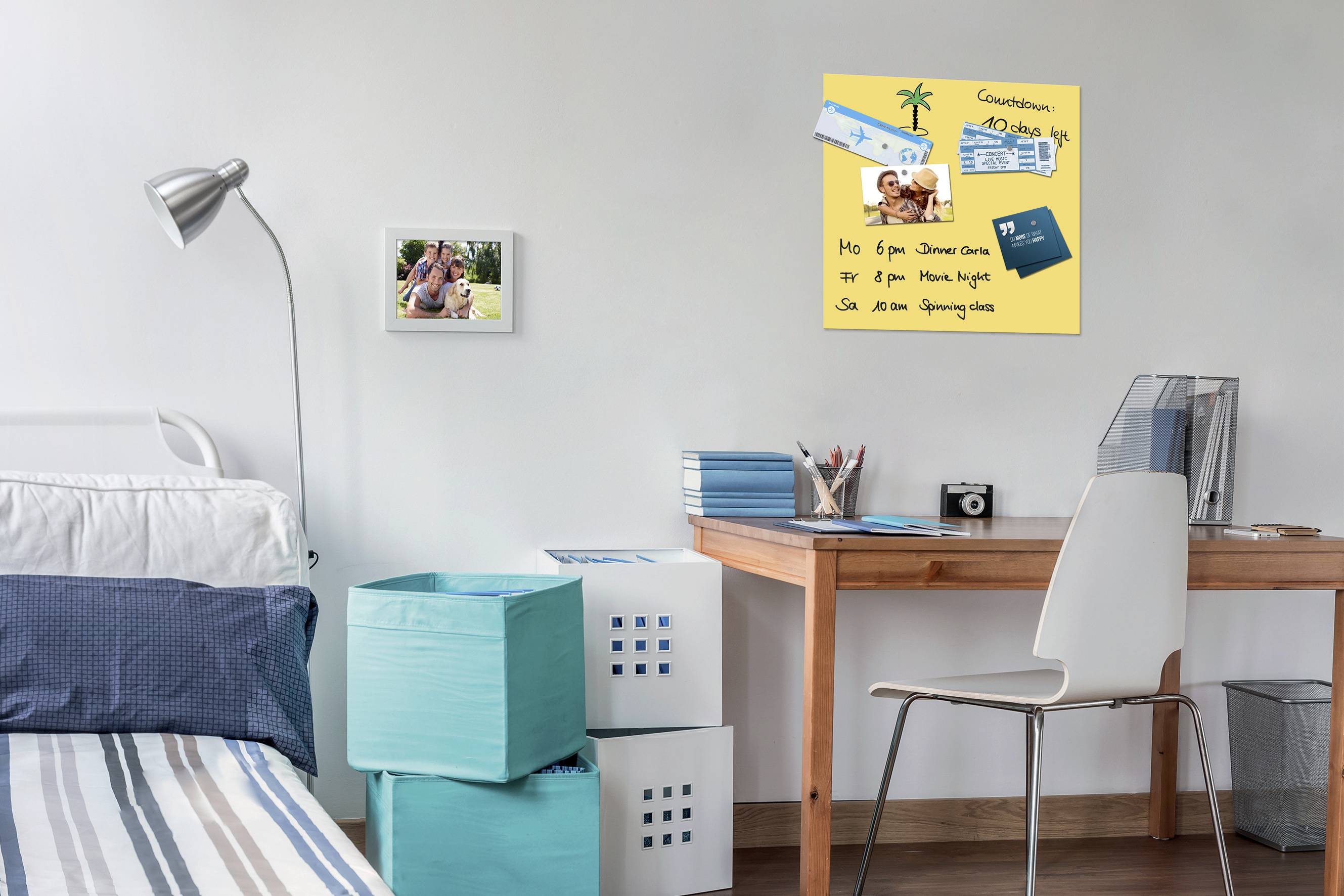 Un tavolo con una sedia è posizionato in una camera da letto. Sulla parete è appesa una bacheca con foto, appunti e un biglietto d'ingresso.