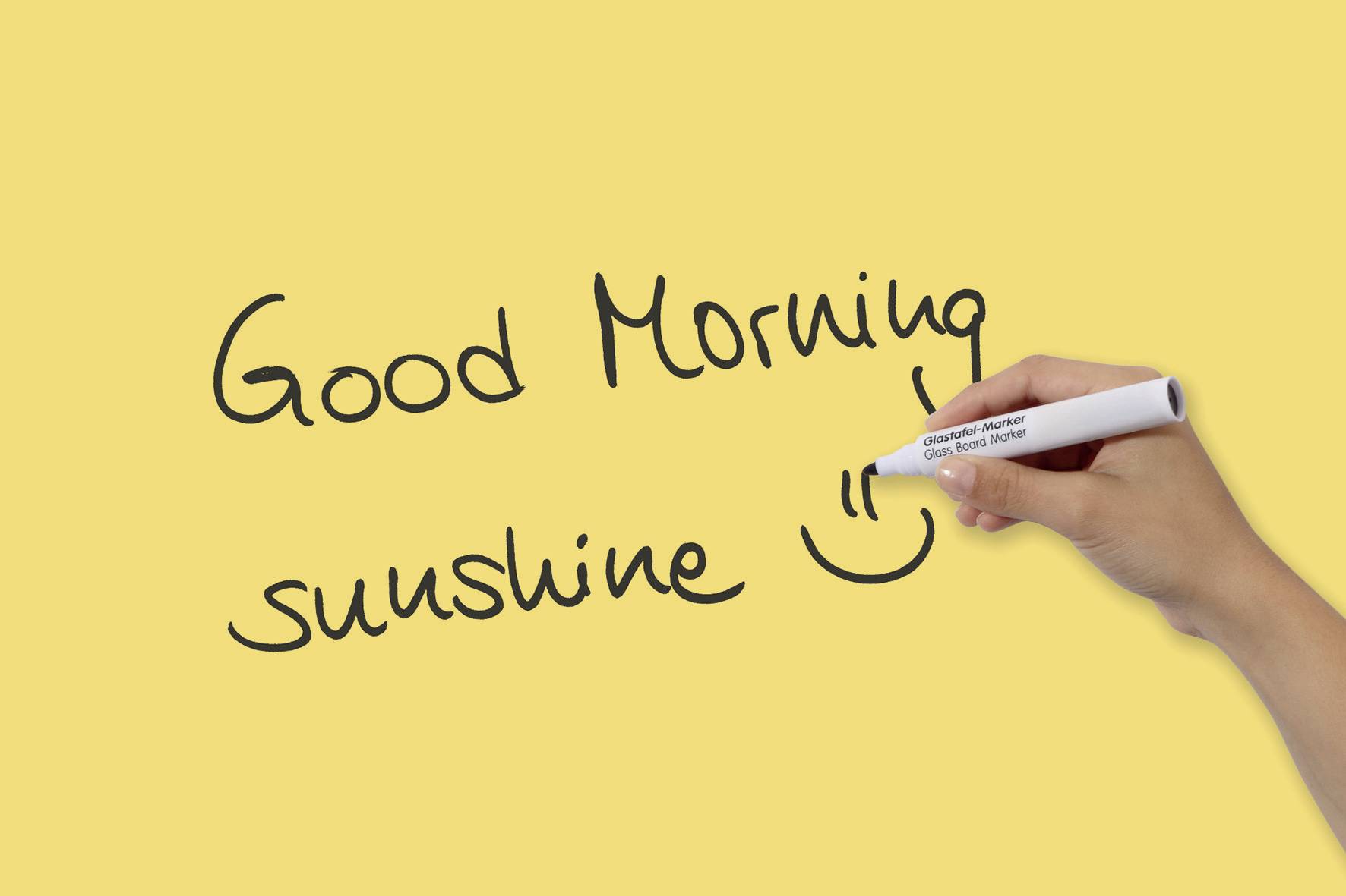 Una mano scrive con una penna bianca su uno sfondo giallo: 'Good Morning sunshine ☺️'.
