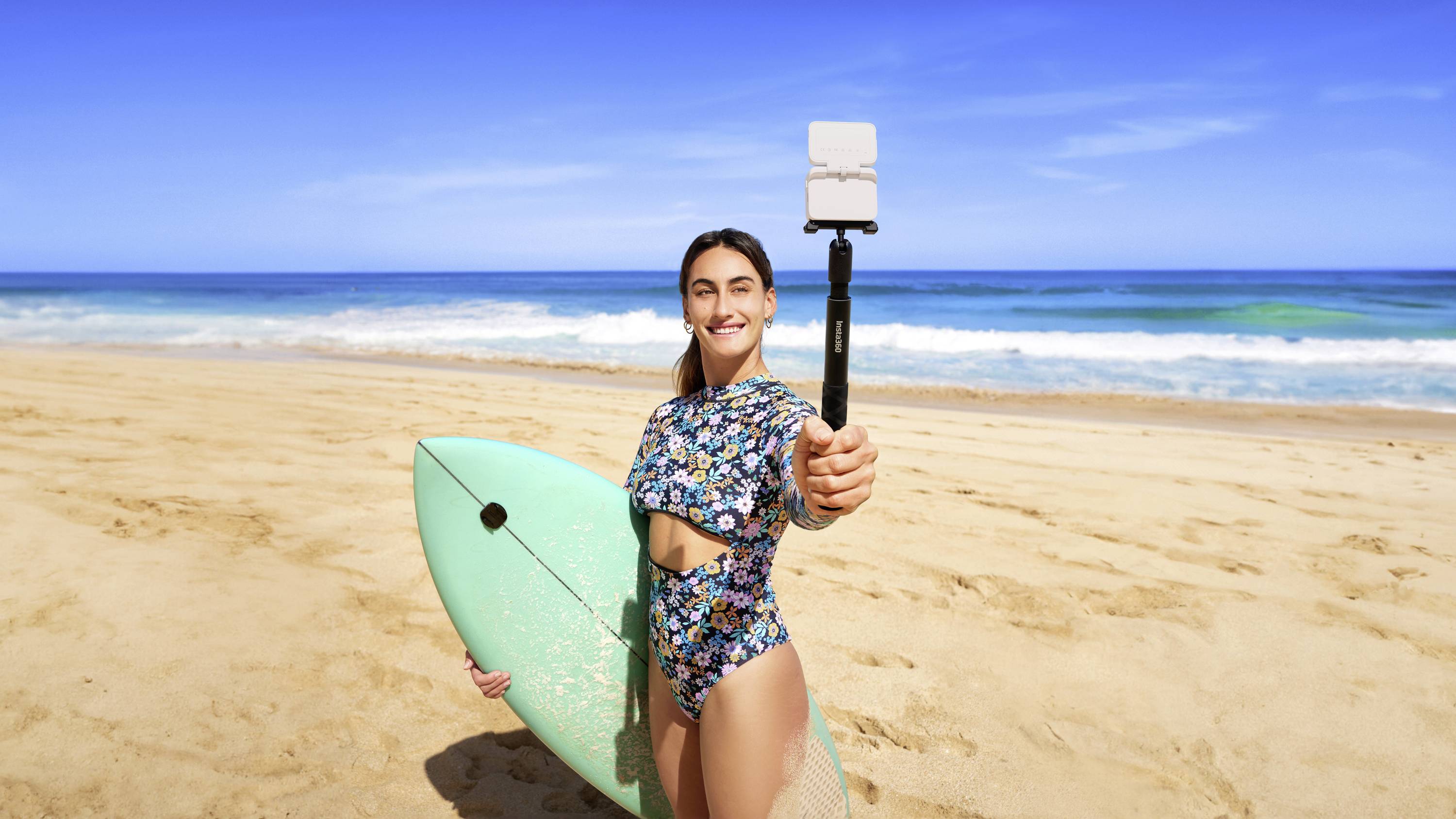 Una persona sorridente sulla spiaggia tiene una tavola da surf e scatta un selfie con un bastone per selfie. Sullo sfondo il mare e il cielo blu.