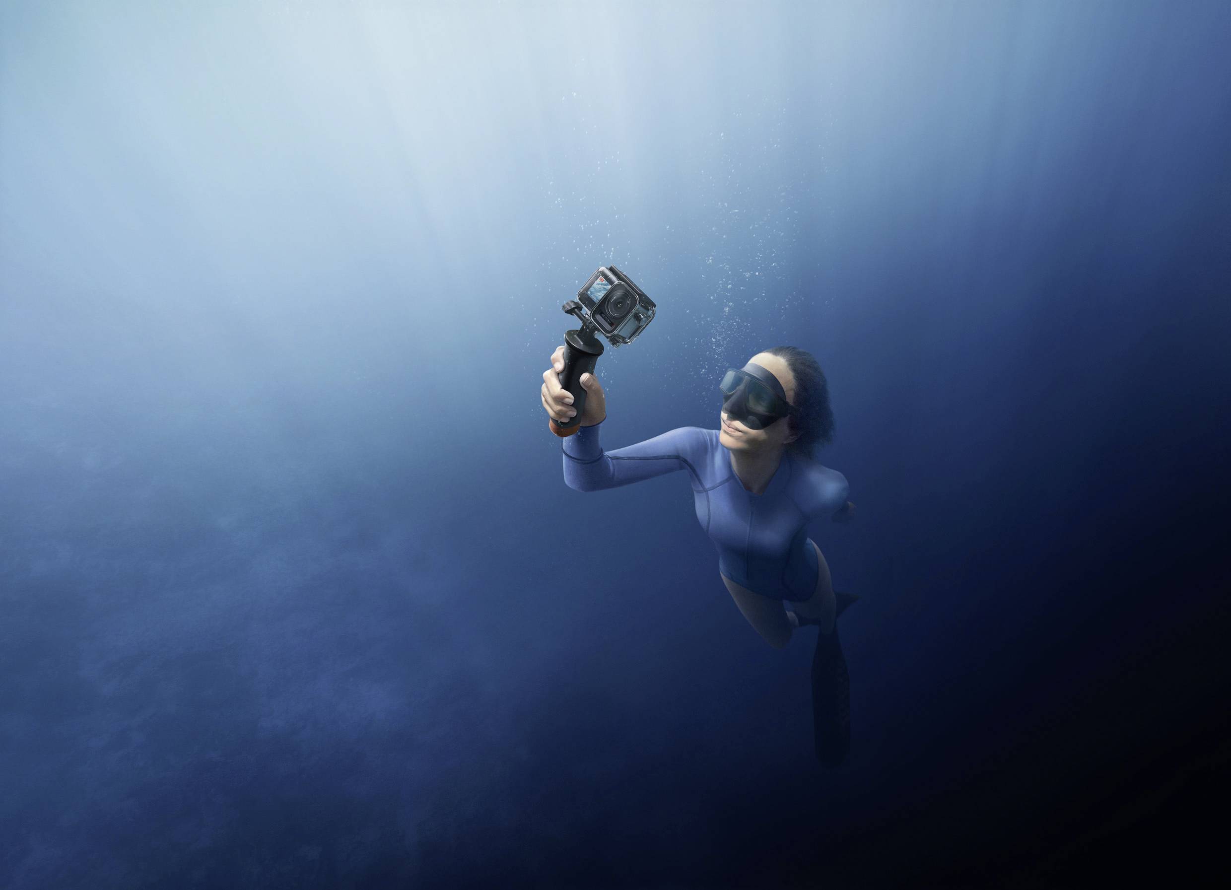 Una persona nuota sott'acqua, tenendo una fotocamera in mano, circondata da luce blu e acqua.