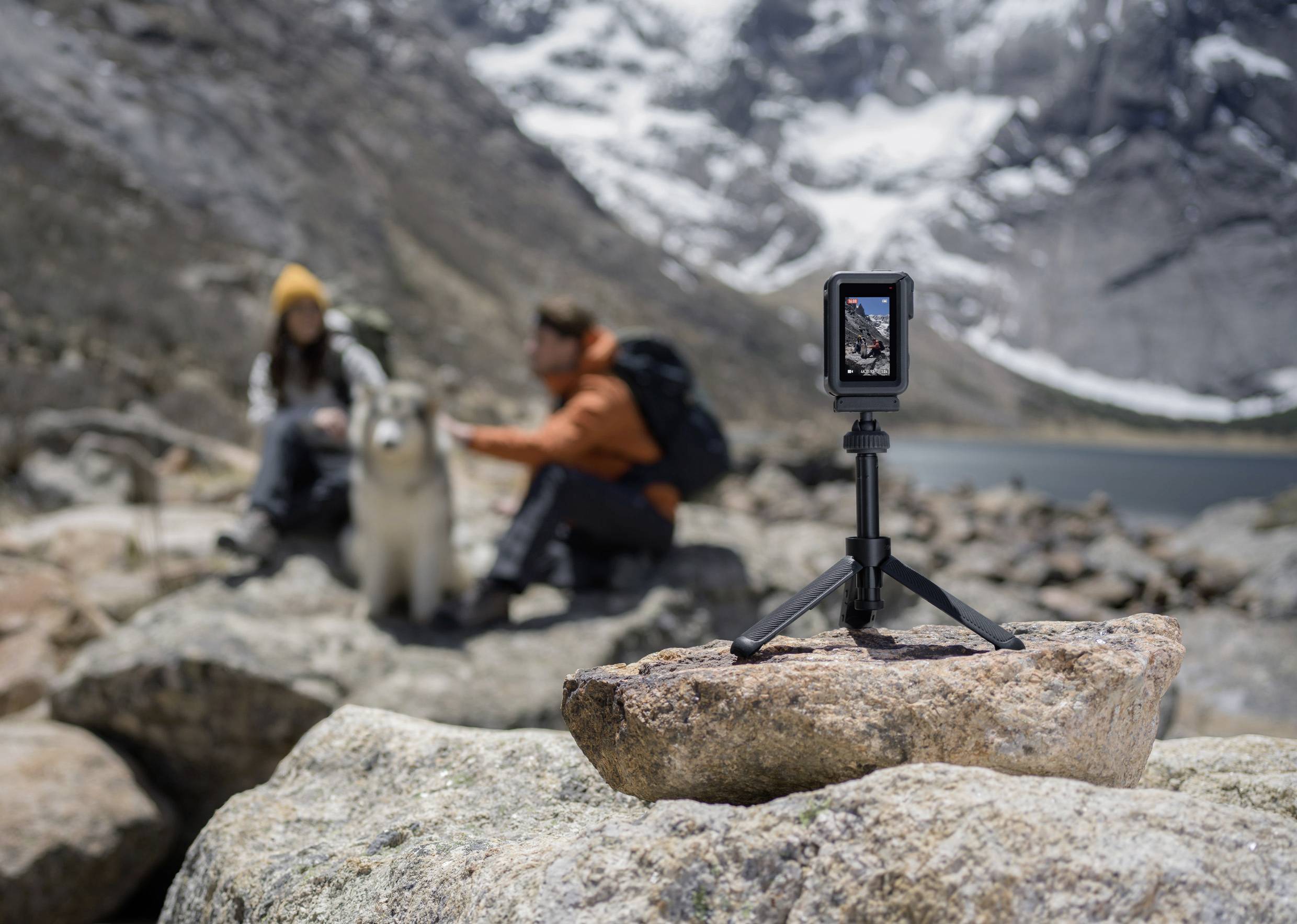 Una fotocamera su un treppiede inquadra una coppia seduta in un paesaggio montano con un cane. Sullo sfondo si vedono montagne innevate.