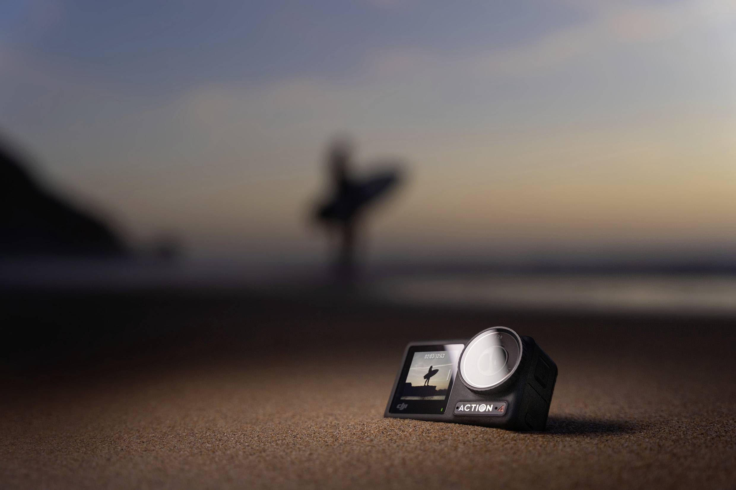 Fotocamera d'azione in primo piano su una spiaggia sabbiosa, sagoma sfocata di un surfista sullo sfondo, atmosfera di tramonto.