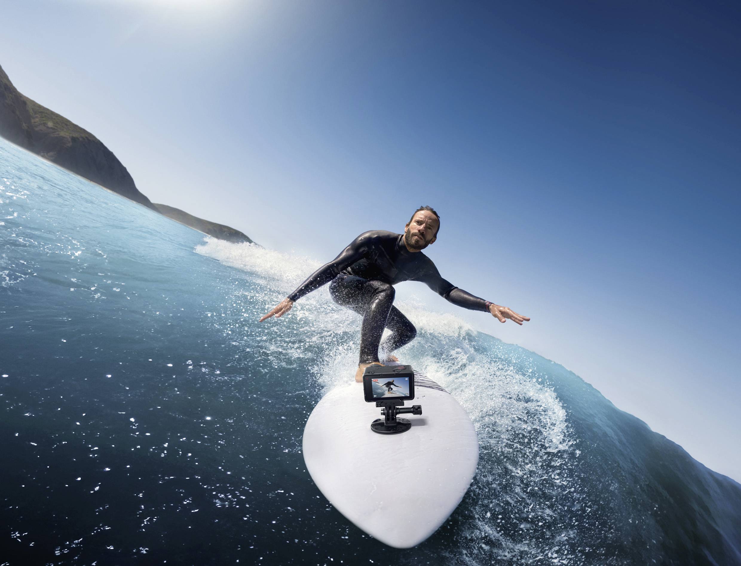 Un surfista cavalca un'onda nel mare con una fotocamera sul suo surf, con un cielo blu e un paesaggio montuoso sullo sfondo.