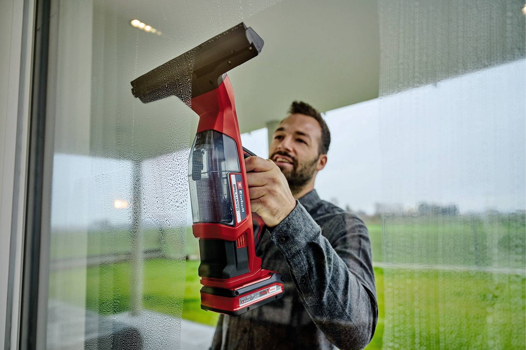 Un uomo sta pulendo accuratamente una grande finestra con un detergente per vetri rosso. Sullo sfondo è visibile un paesaggio verde.