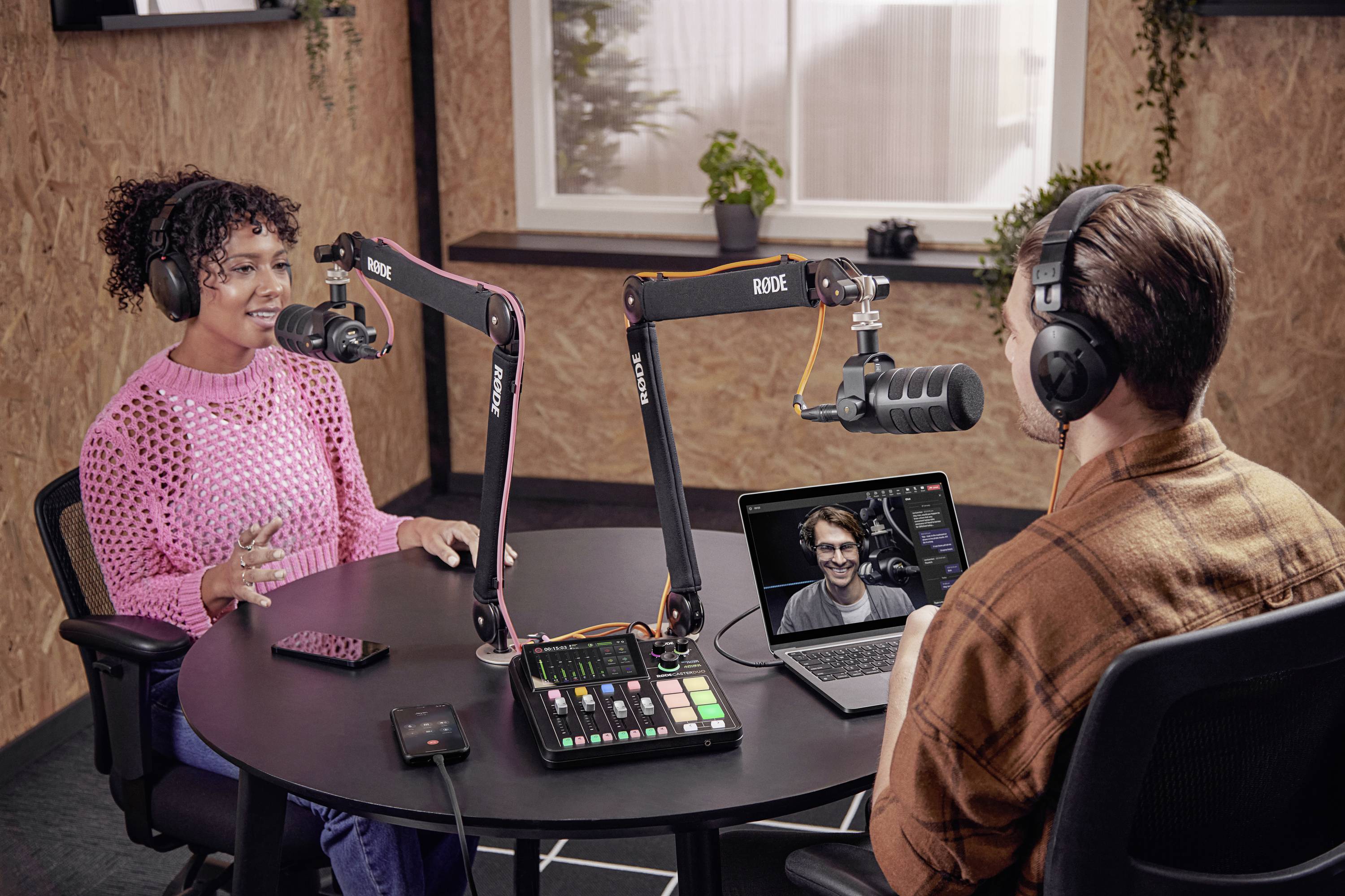 Donna e uomo con le cuffie registrano un podcast in uno studio. Un laptop mostra una videoconferenza con un'altra persona.