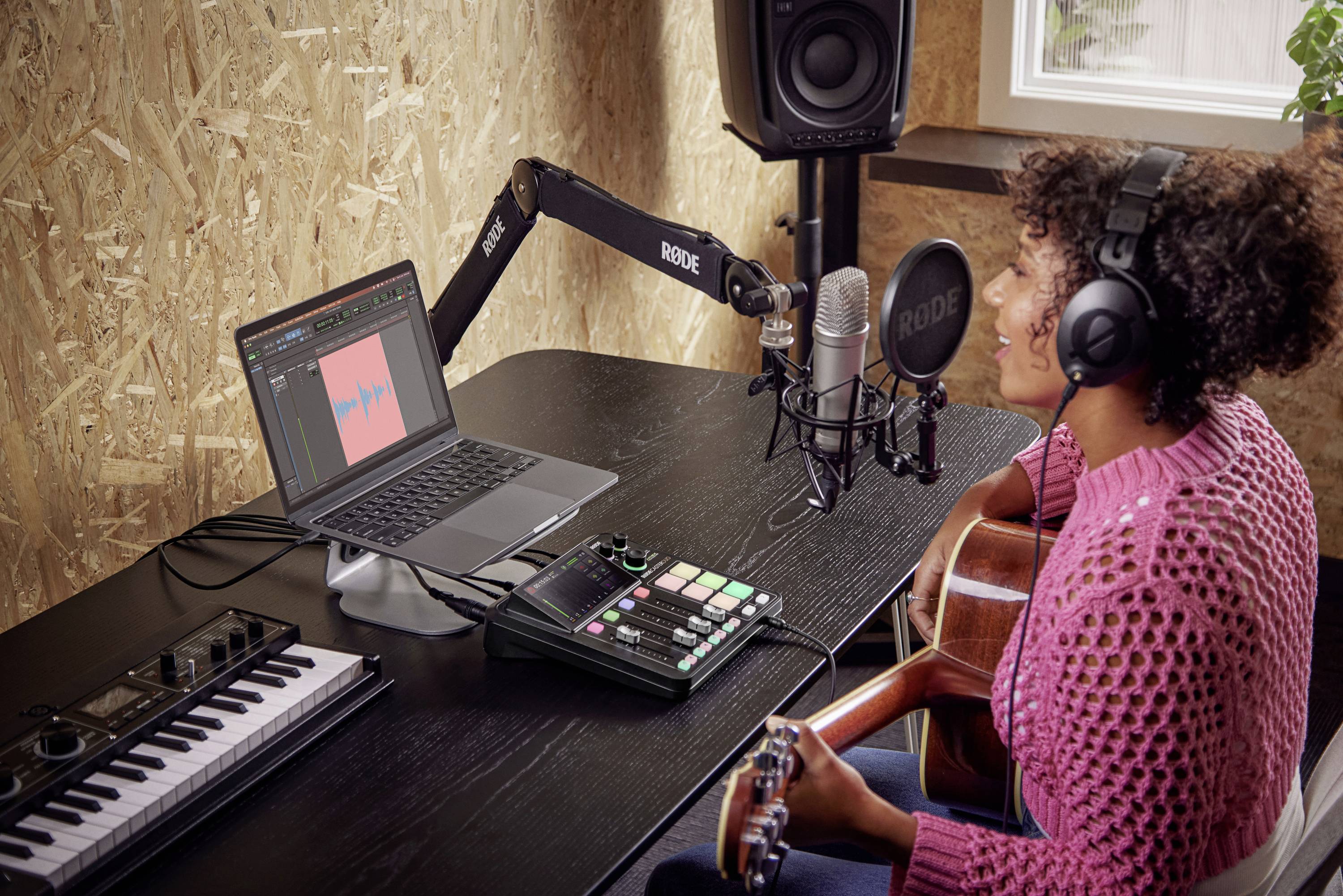 Una persona con le cuffie suona la chitarra e registra musica. Davanti a sé ha un laptop con software audio, microfono e controller MIDI.