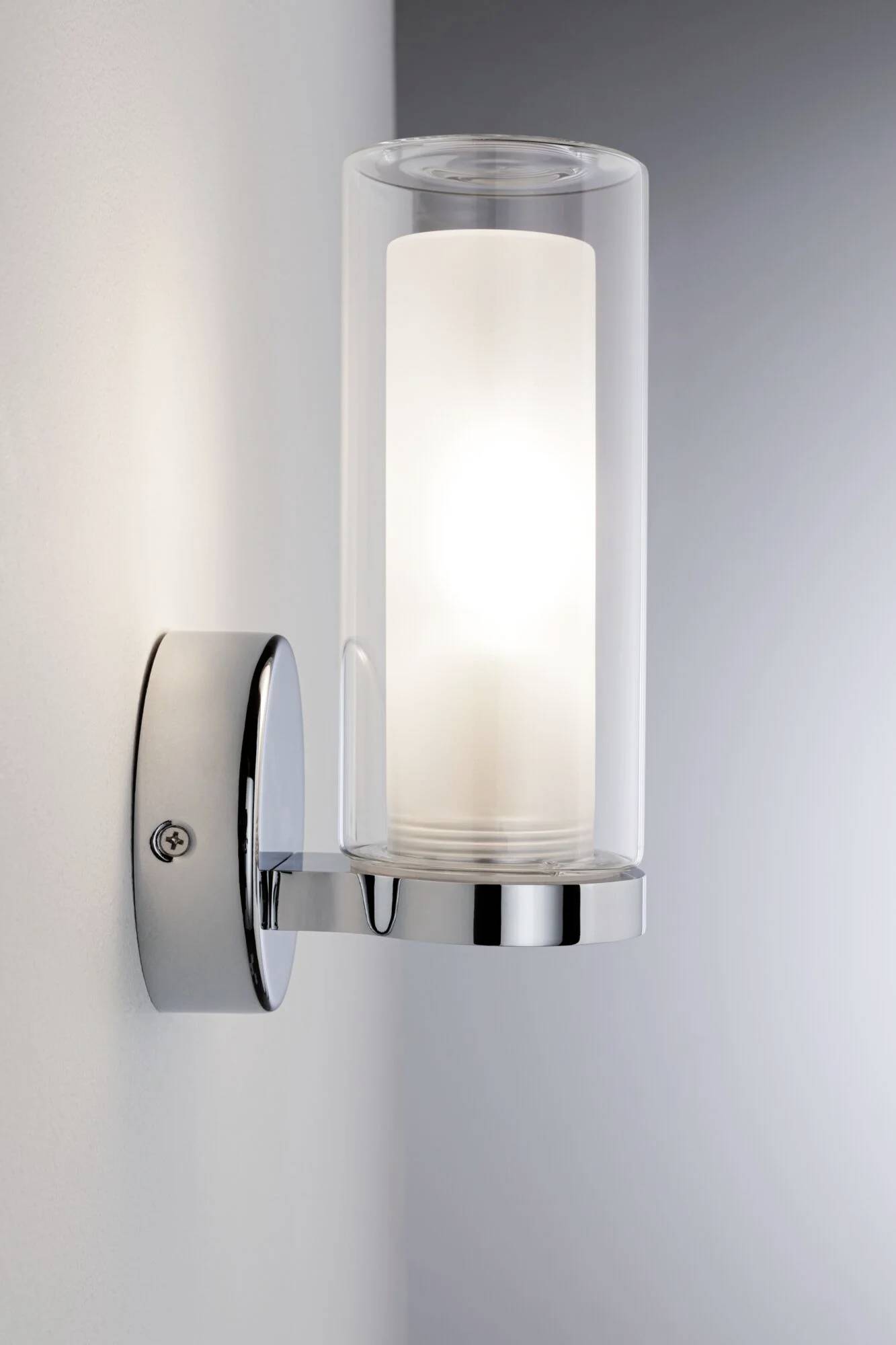 Lampada da parete moderna con vetro cilindrico e supporto cromato, che emana una luce calda.