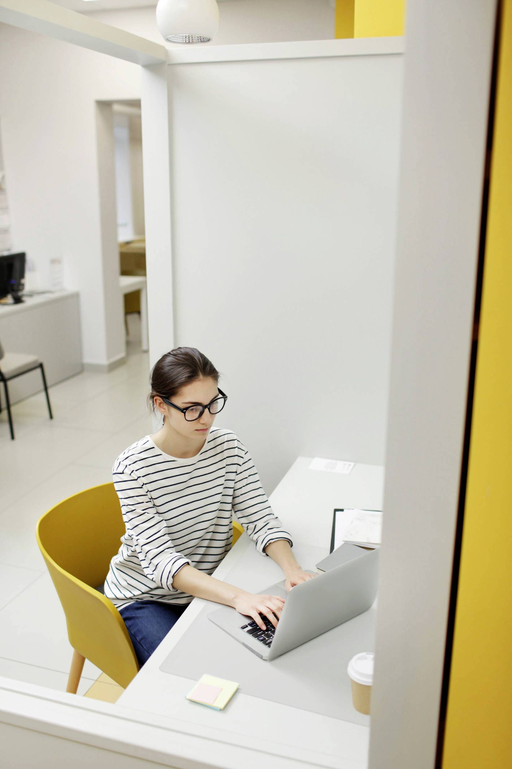 Una donna con gli occhiali lavora su un laptop in un ufficio, seduta su una sedia gialla con una tazza di caffè sul tavolo.
