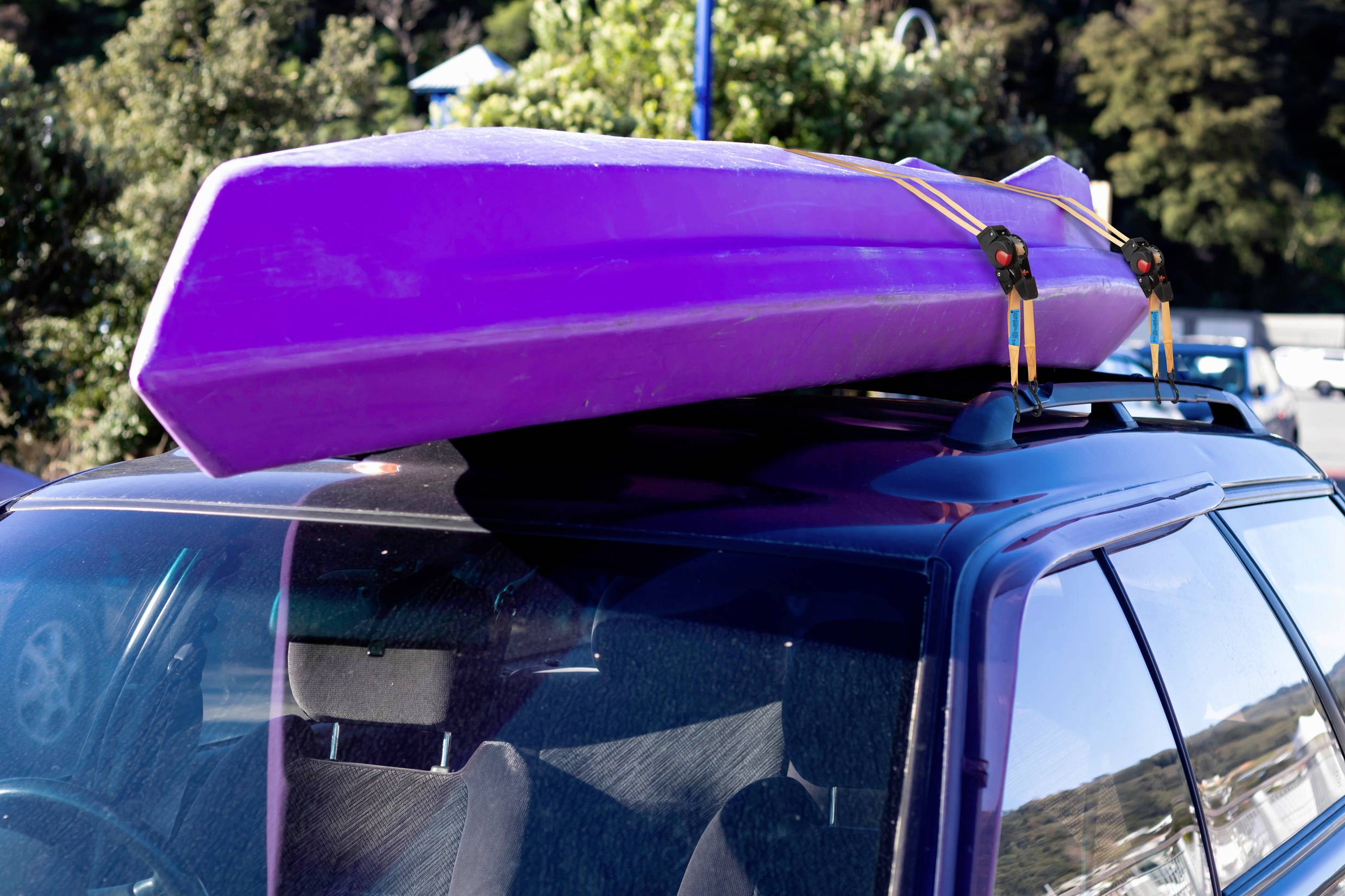 Un kayak lilla è saldamente fissato con cinghie sul tetto di un'auto. Sullo sfondo si vedono degli alberi.