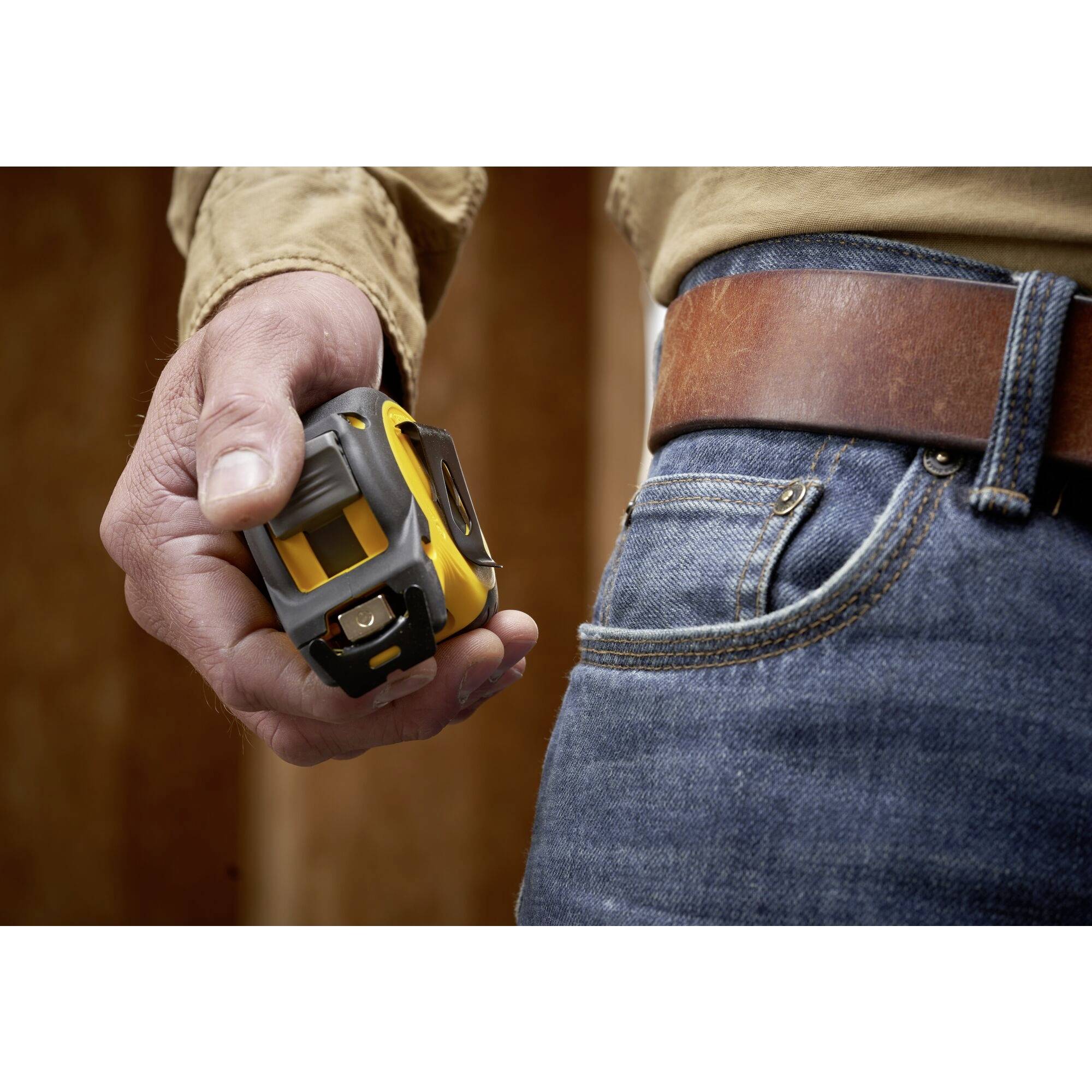 Una persona tiene in mano un nastro metrico giallo. Indossa jeans con una cintura in pelle marrone, visibile sullo sfondo di legno.