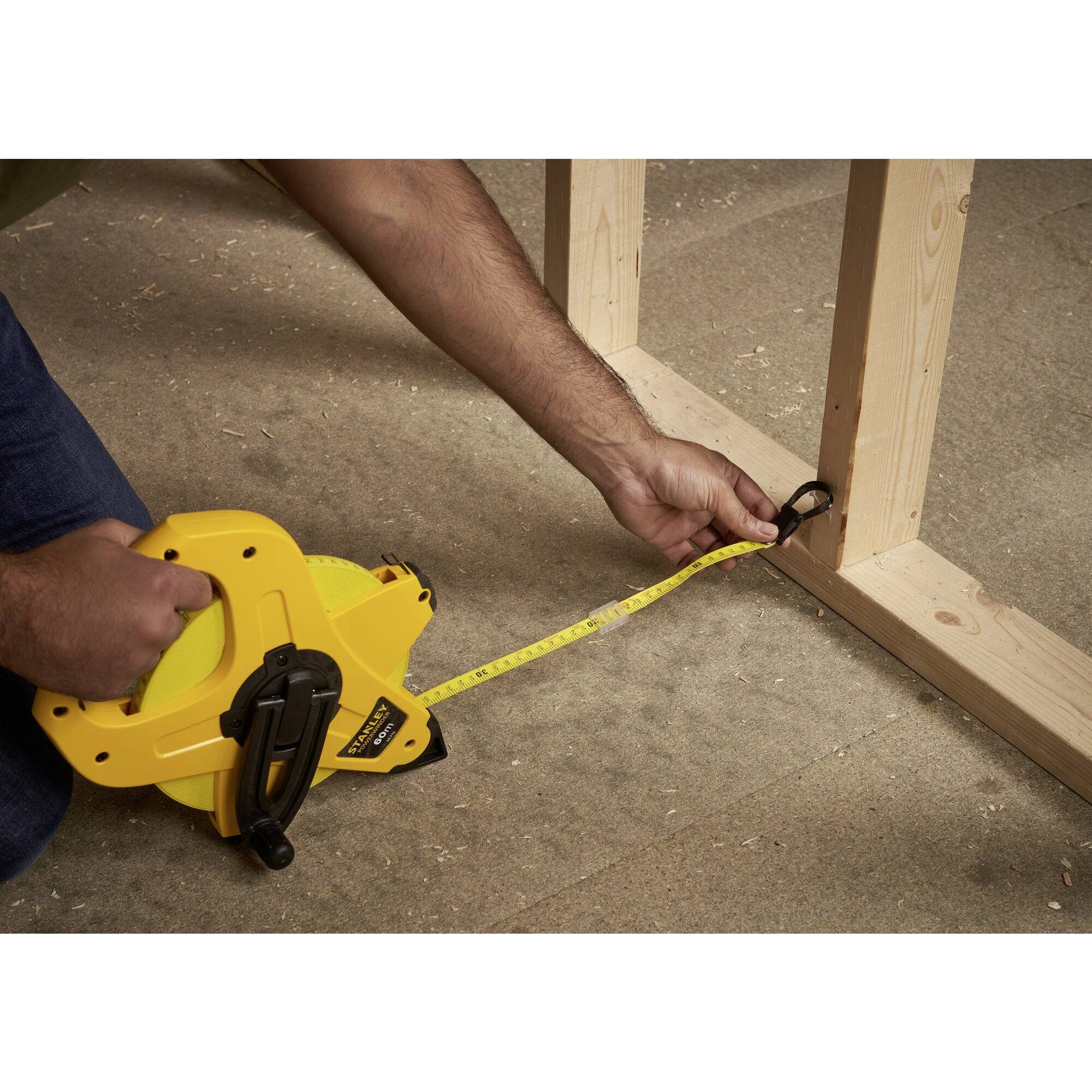Una persona misura la distanza tra cornici di legno su un pavimento rivestito di moquette utilizzando un nastro metrico giallo.