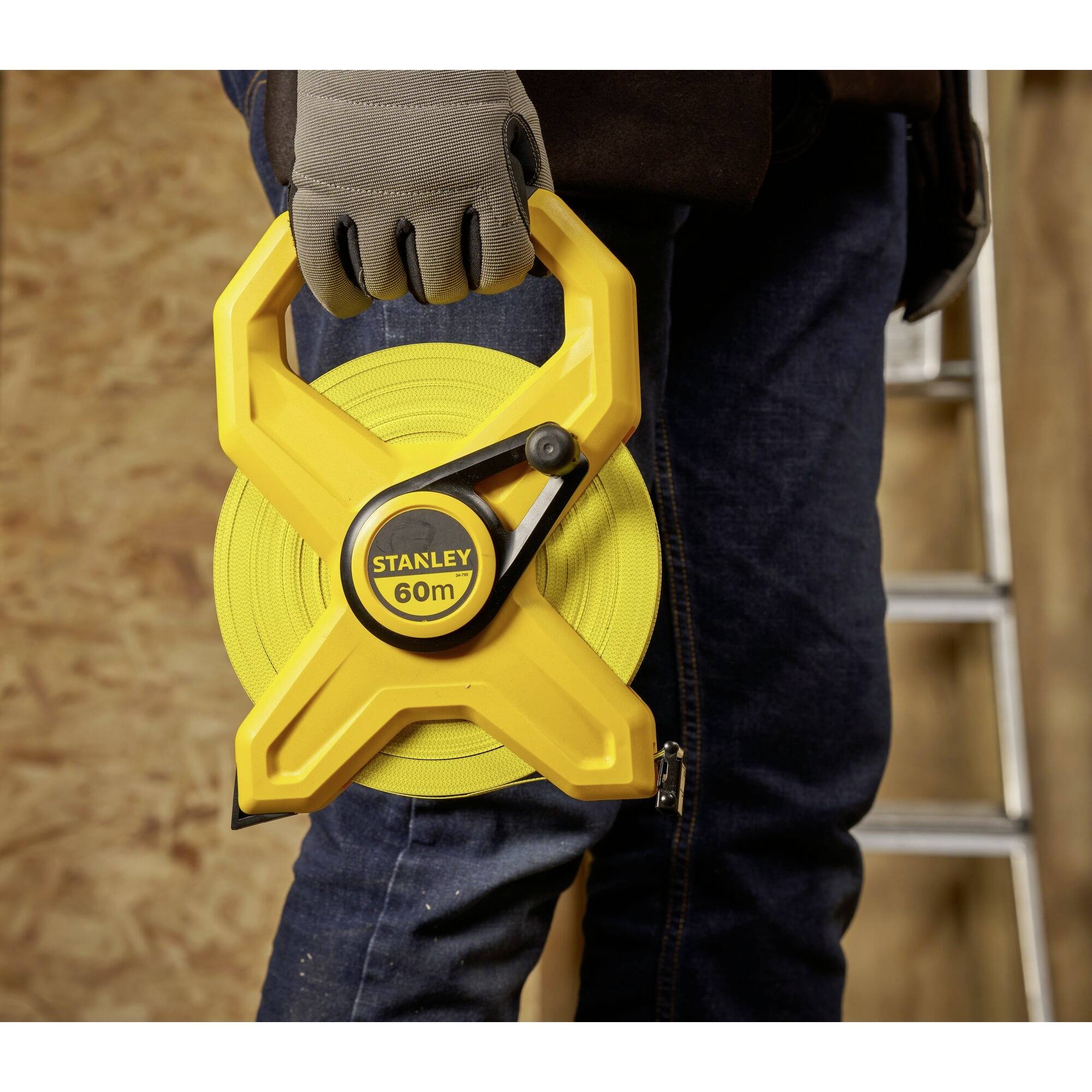 Un lavoratore edile tiene in mano un nastro metrico giallo con la scritta 'STANLEY 60m'. Sullo sfondo si vedono una tavola di legno e una scala.