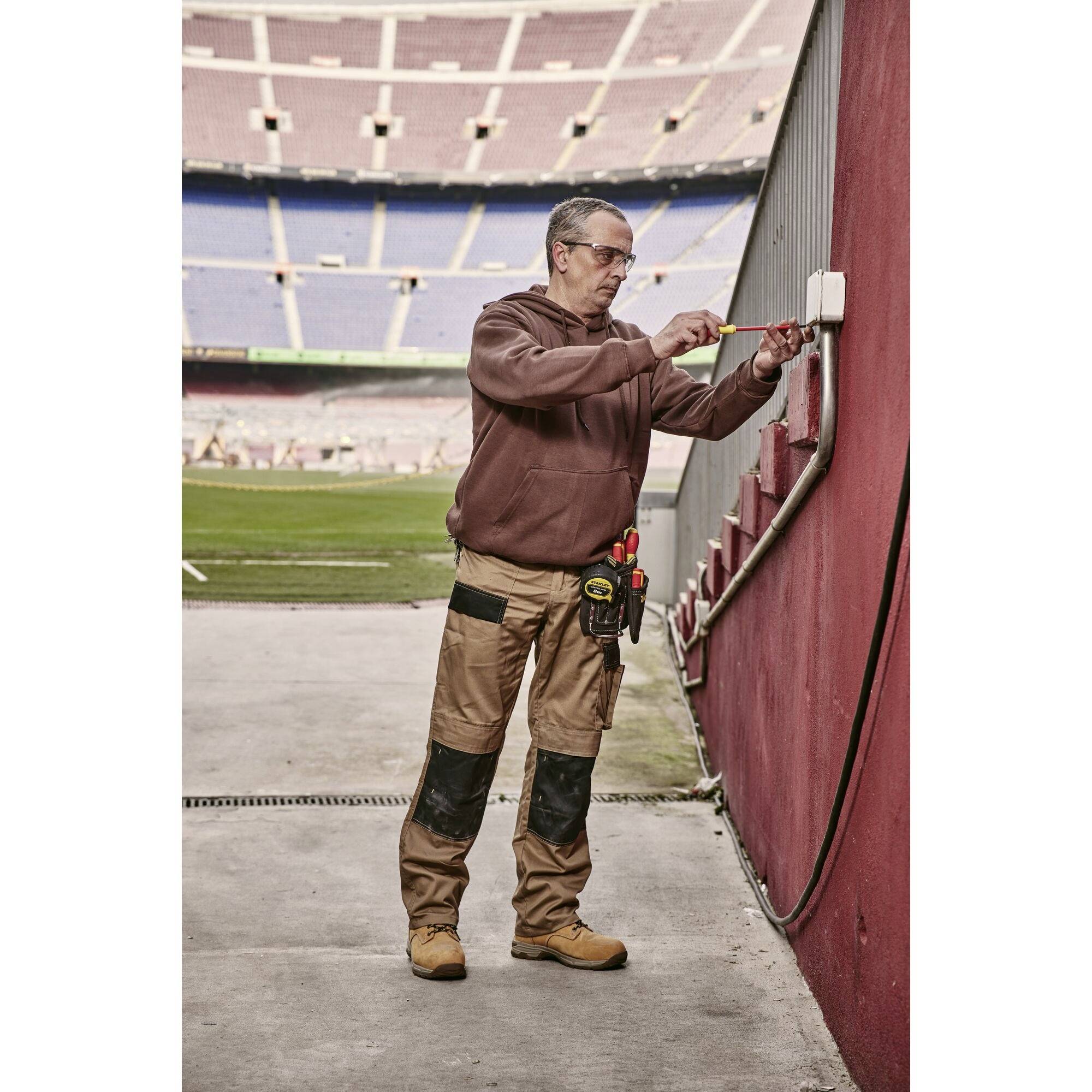 Un operaio in uno stadio sta riparando un cavo elettrico su una parete. Indossa abiti da lavoro e sta eseguendo interventi di manutenzione.