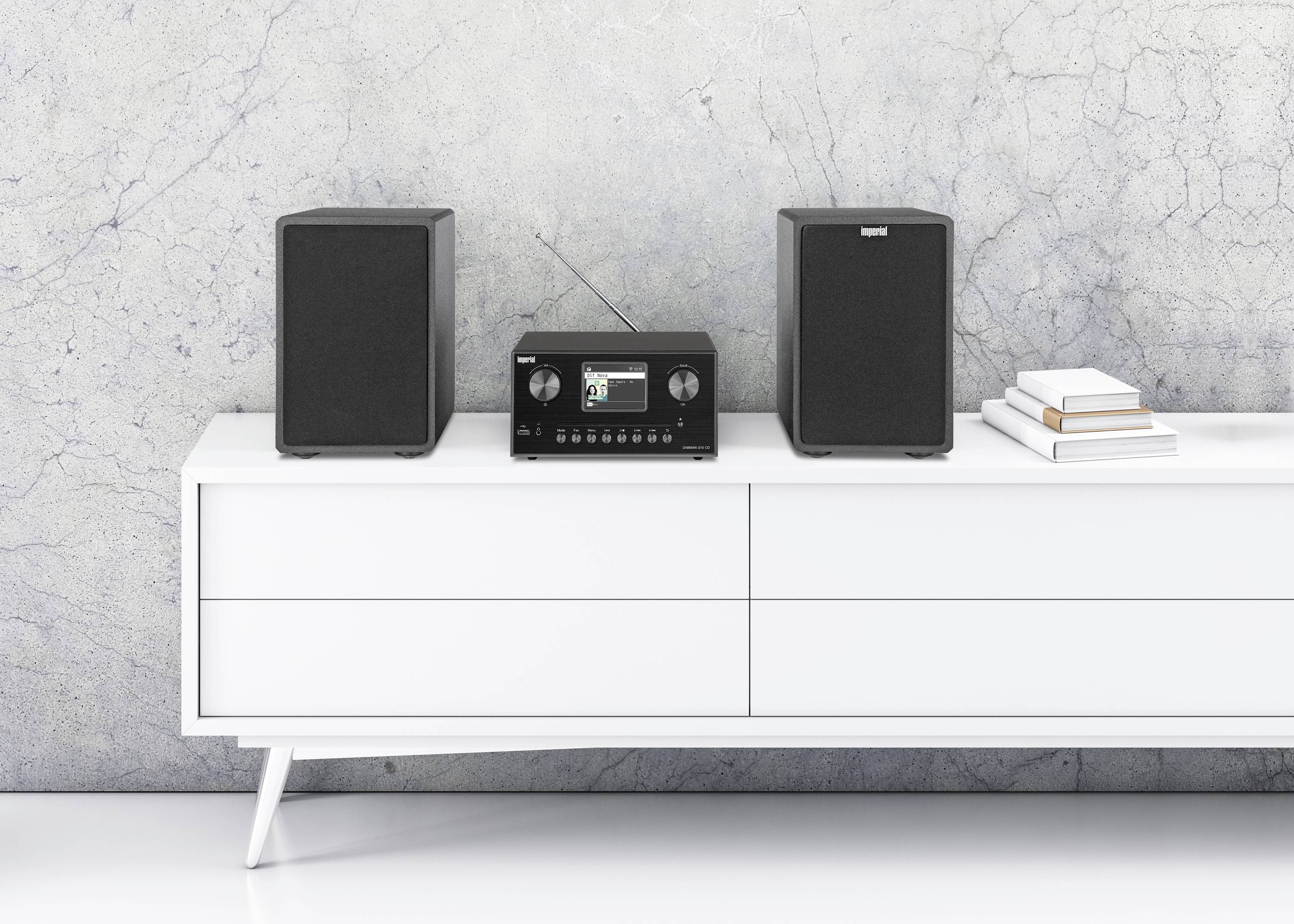 Un impianto stereo con due altoparlanti è posizionato su un mobile bianco davanti a una parete grigia con texture. Dei libri sono appoggiati a destra.