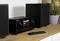 Impianto stereo nero con display digitale e due altoparlanti su un tavolo di legno, accanto un telecomando e piante sullo sfondo.