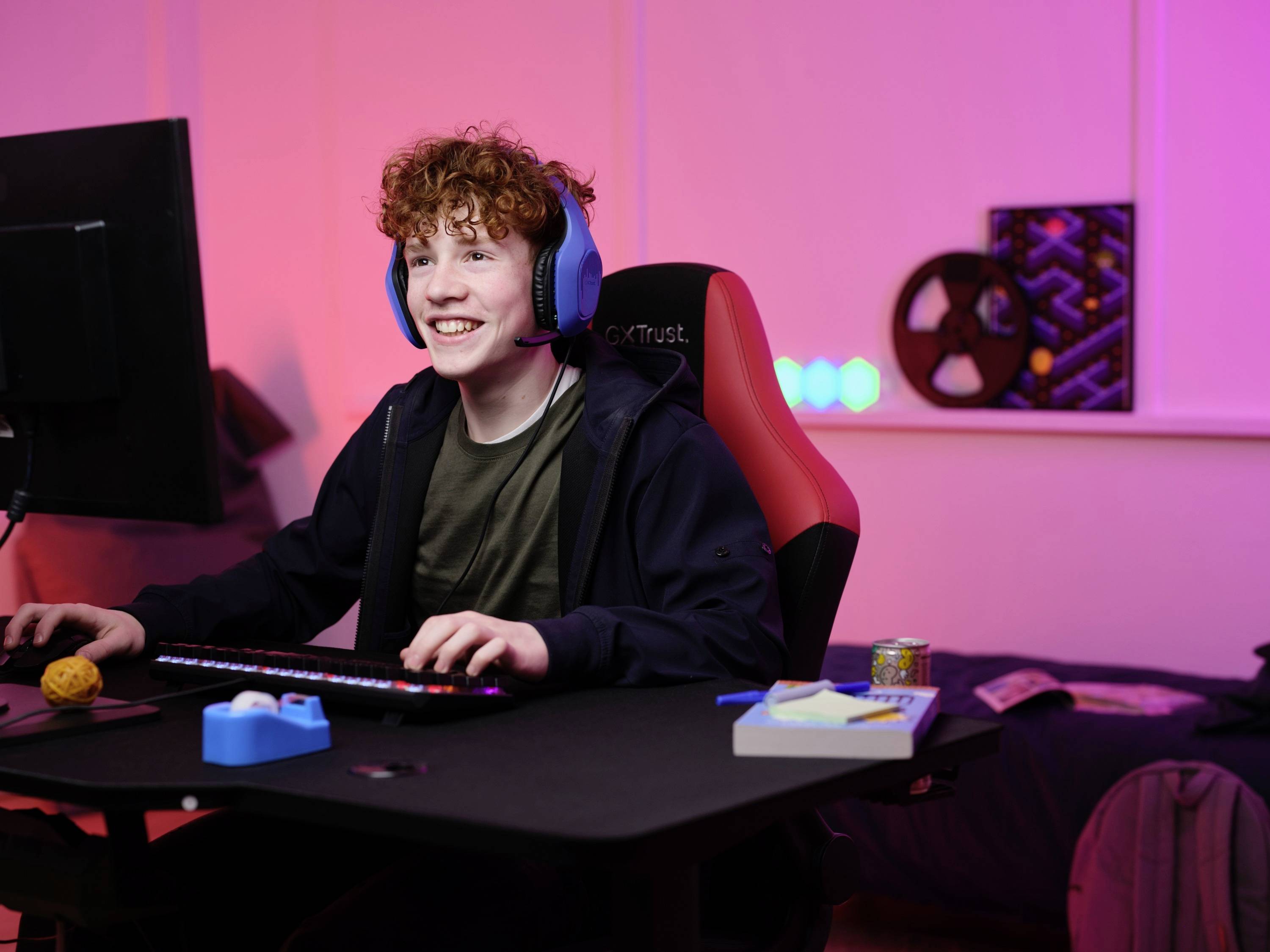 Un adolescente sorride seduto davanti a un PC, indossando delle cuffie, in una stanza arredata in modo moderno con un'illuminazione colorata.