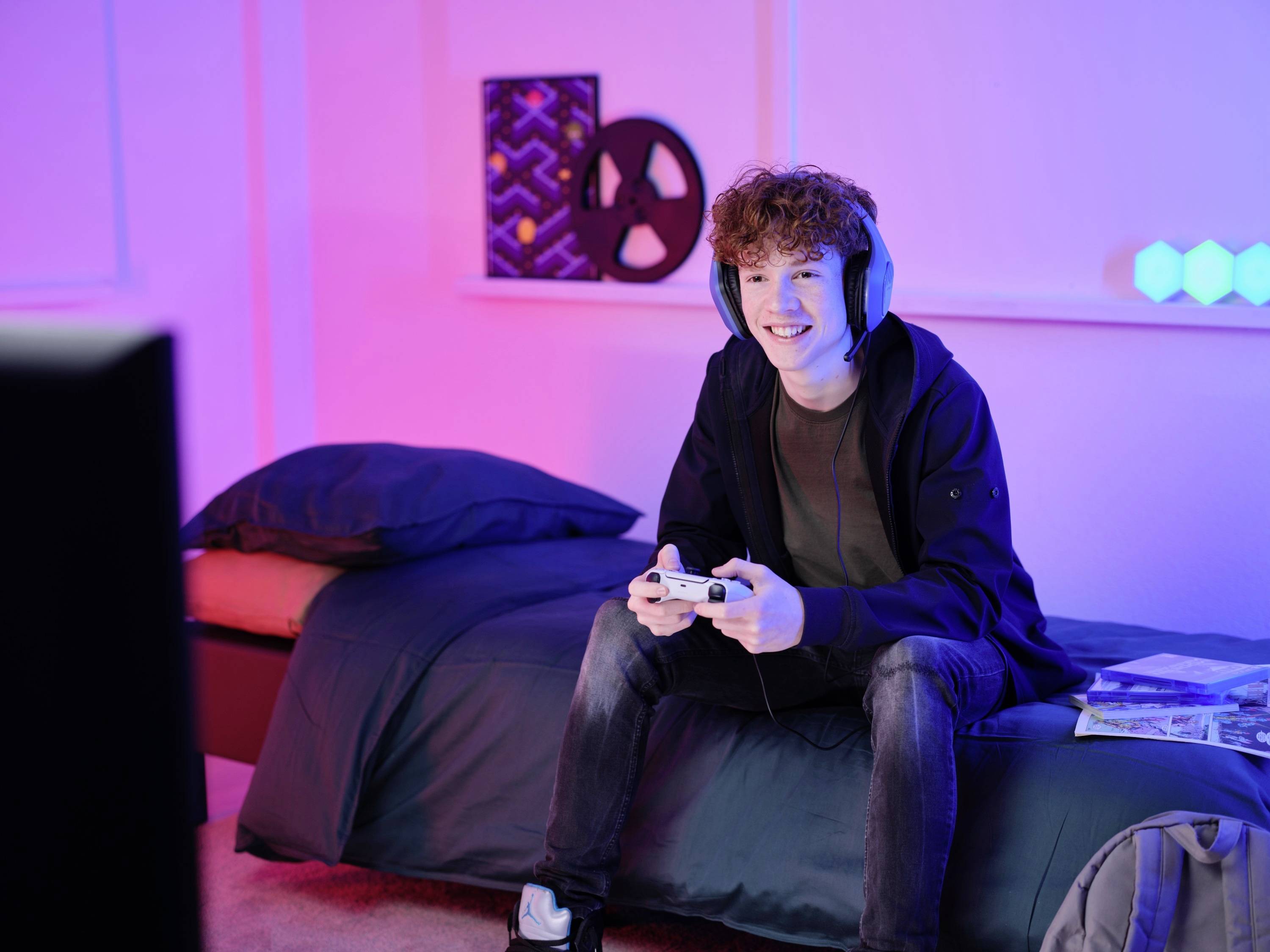 Giovane uomo seduto su un letto con le cuffie, intento a giocare con un controller. Sullo sfondo, luci colorate e un elemento decorativo.