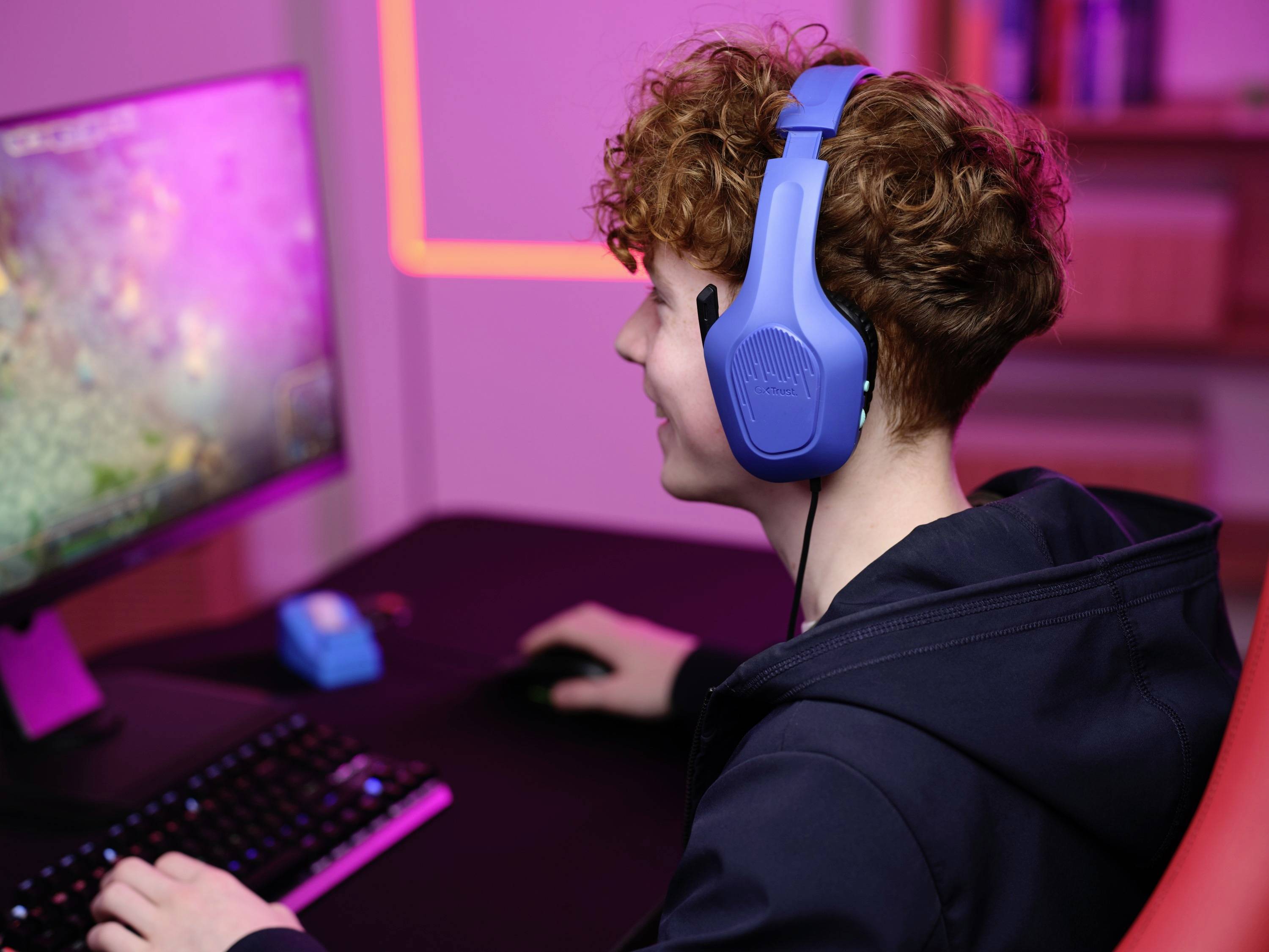 Una persona con cuffie blu seduta a una scrivania davanti a un monitor, mentre digita su una tastiera, avvolta in una luce rosa.