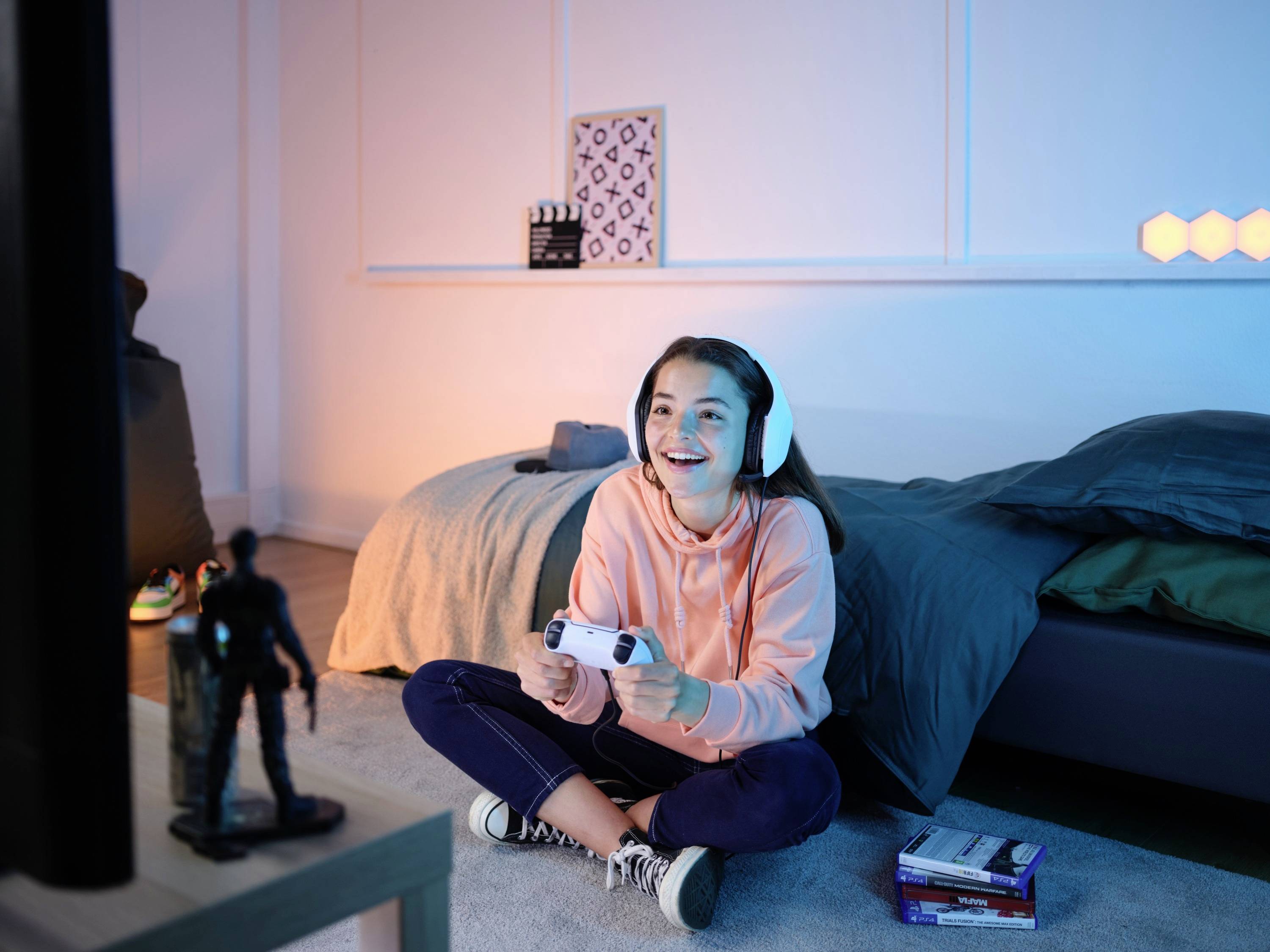 Una ragazza è seduta sul pavimento in una stanza, indossa delle cuffie e gioca con un controller per videogiochi. Sembra felice e concentrata.