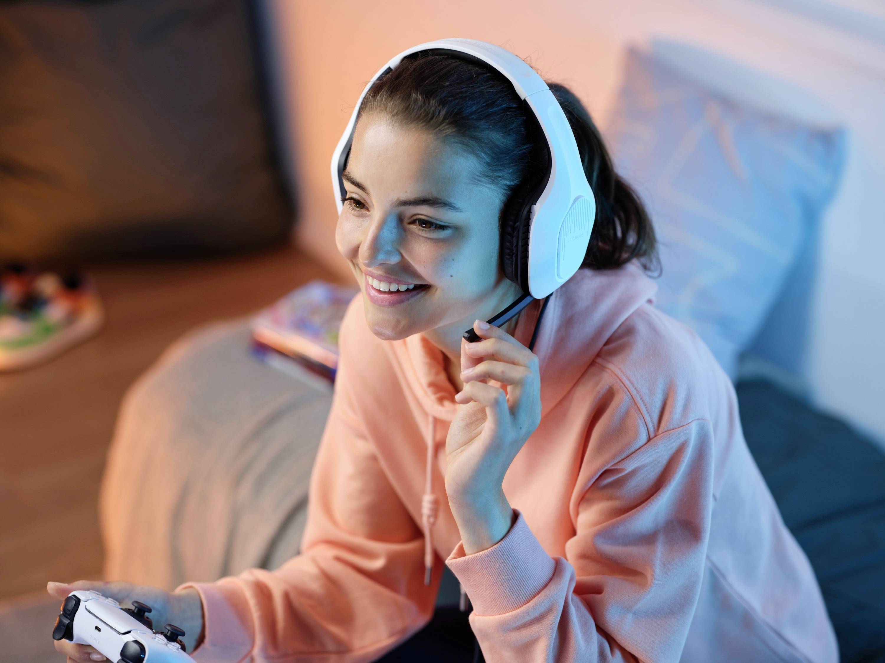 Una persona sorridente con un felpa rosa seduta su un letto, indossa cuffie bianche e tiene in mano un controller per videogiochi.