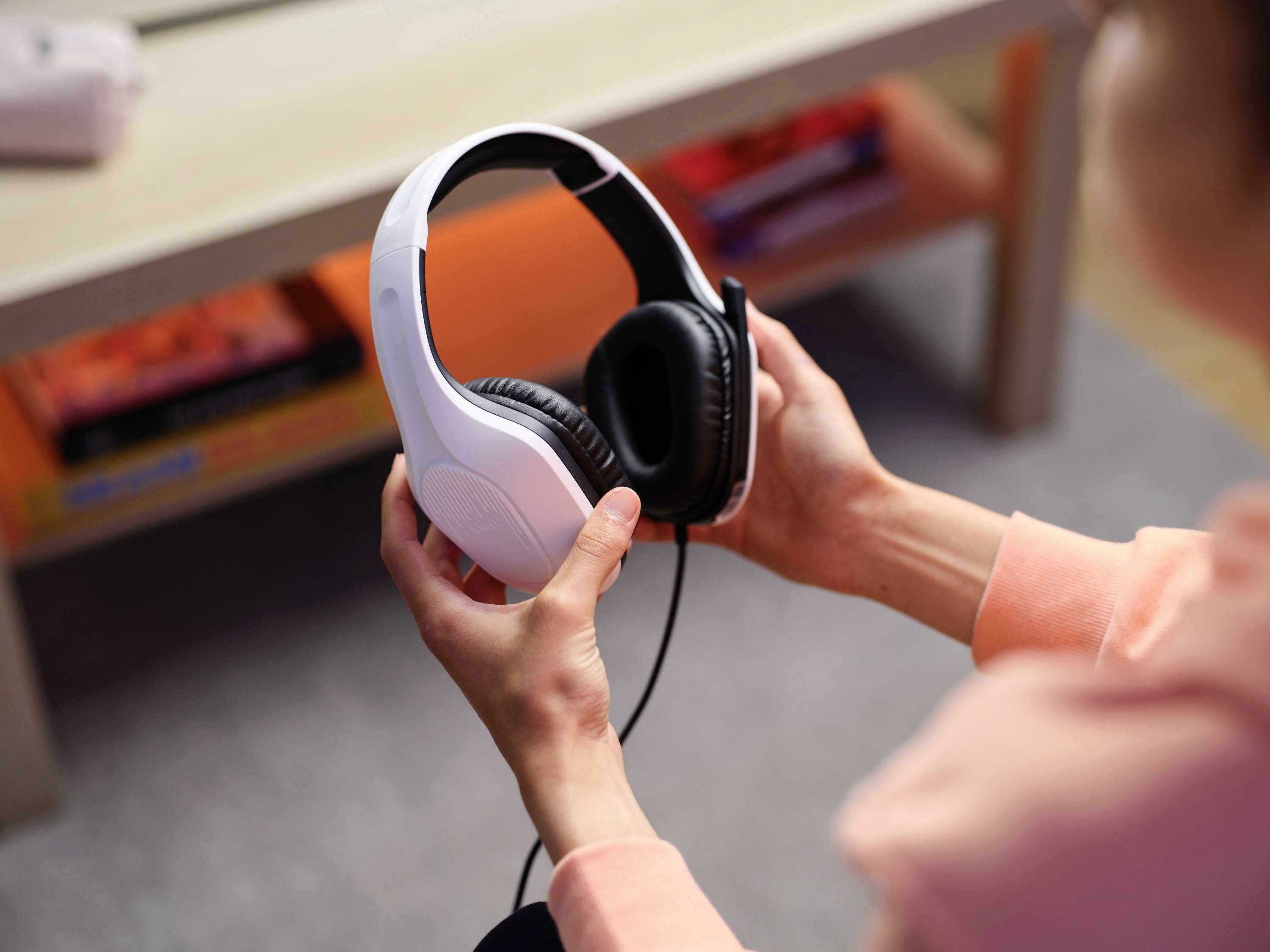 Una persona tiene tra le mani un paio di cuffie over-ear bianche con cavo. Sullo sfondo si trova un tavolo con dei libri.