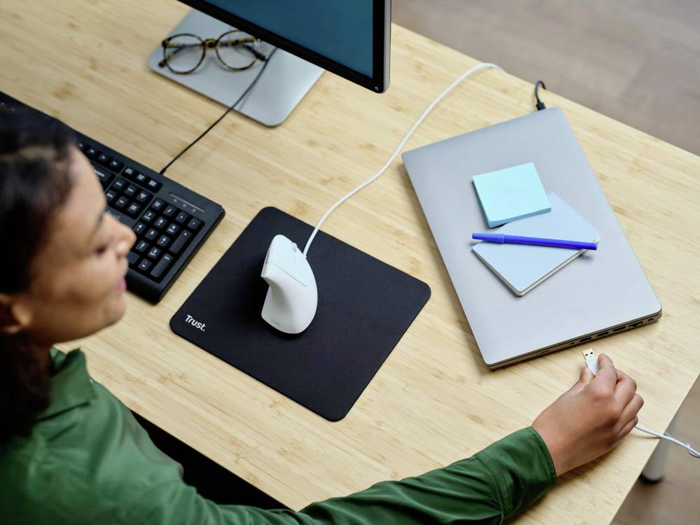 Una persona lavora a una scrivania con un computer, un mouse ergonomico e un laptop. Appunti e una penna sono posizionati sul laptop.