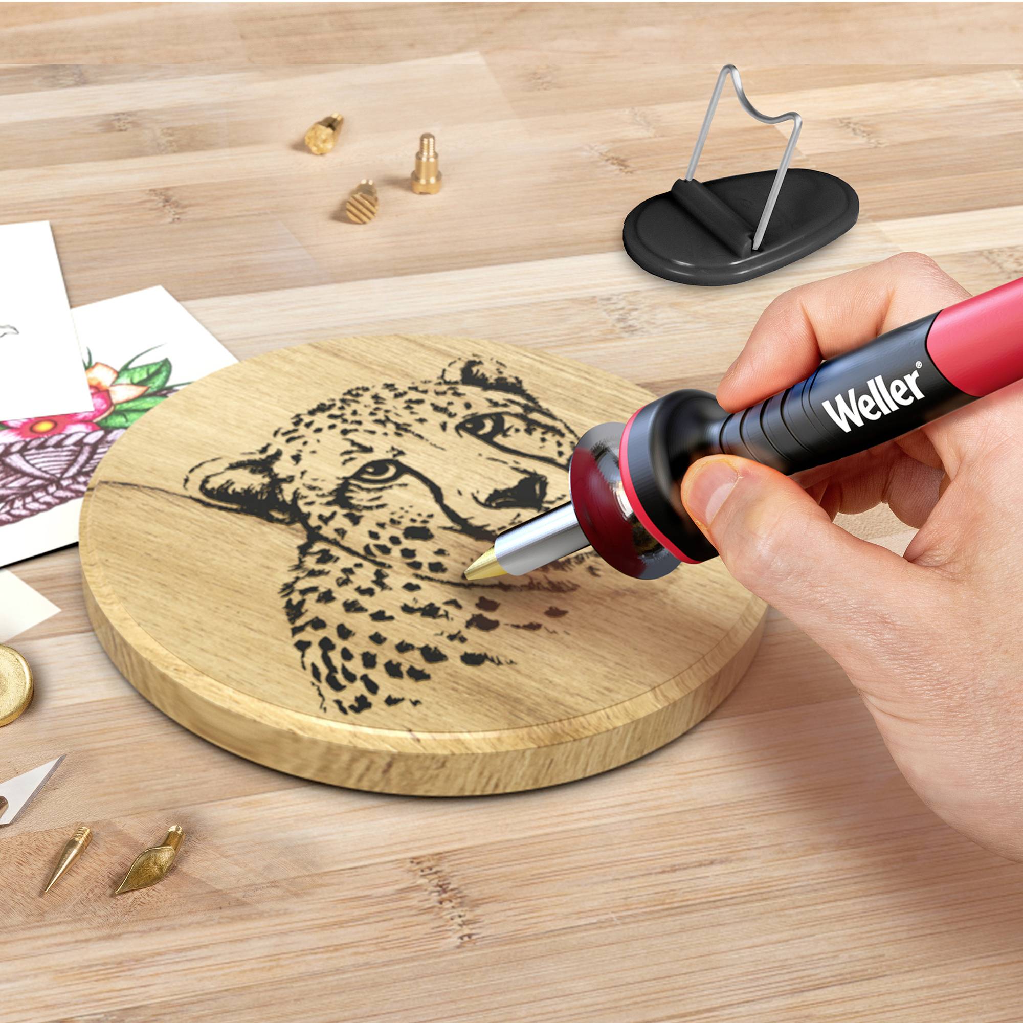 Una mano sta usando un saldatore per incidere l'immagine di un leopardo su una tavola rotonda di legno. Sullo sfondo sono presenti alcuni disegni.