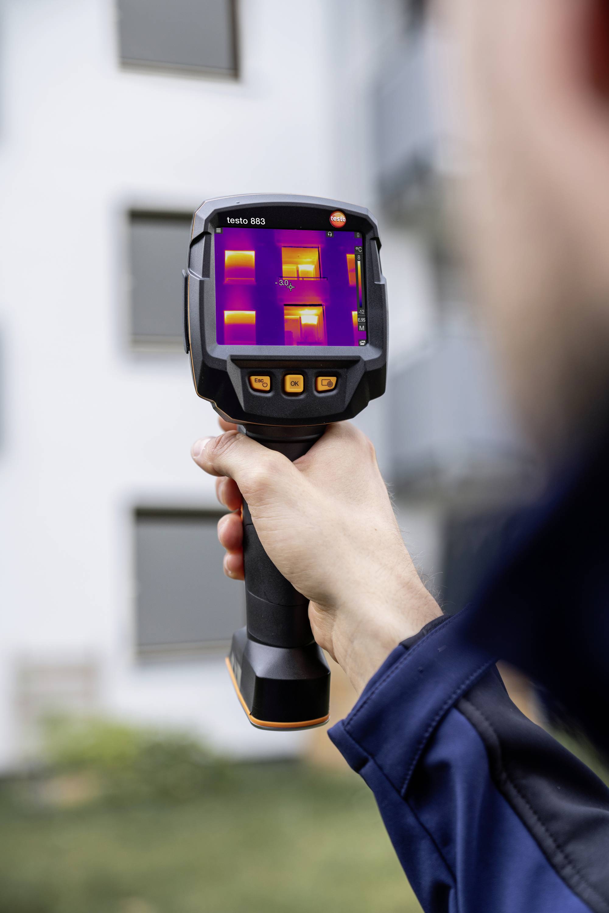 Una persona tiene una termocamera, mostrando un edificio con differenze di temperatura visibili sul display.