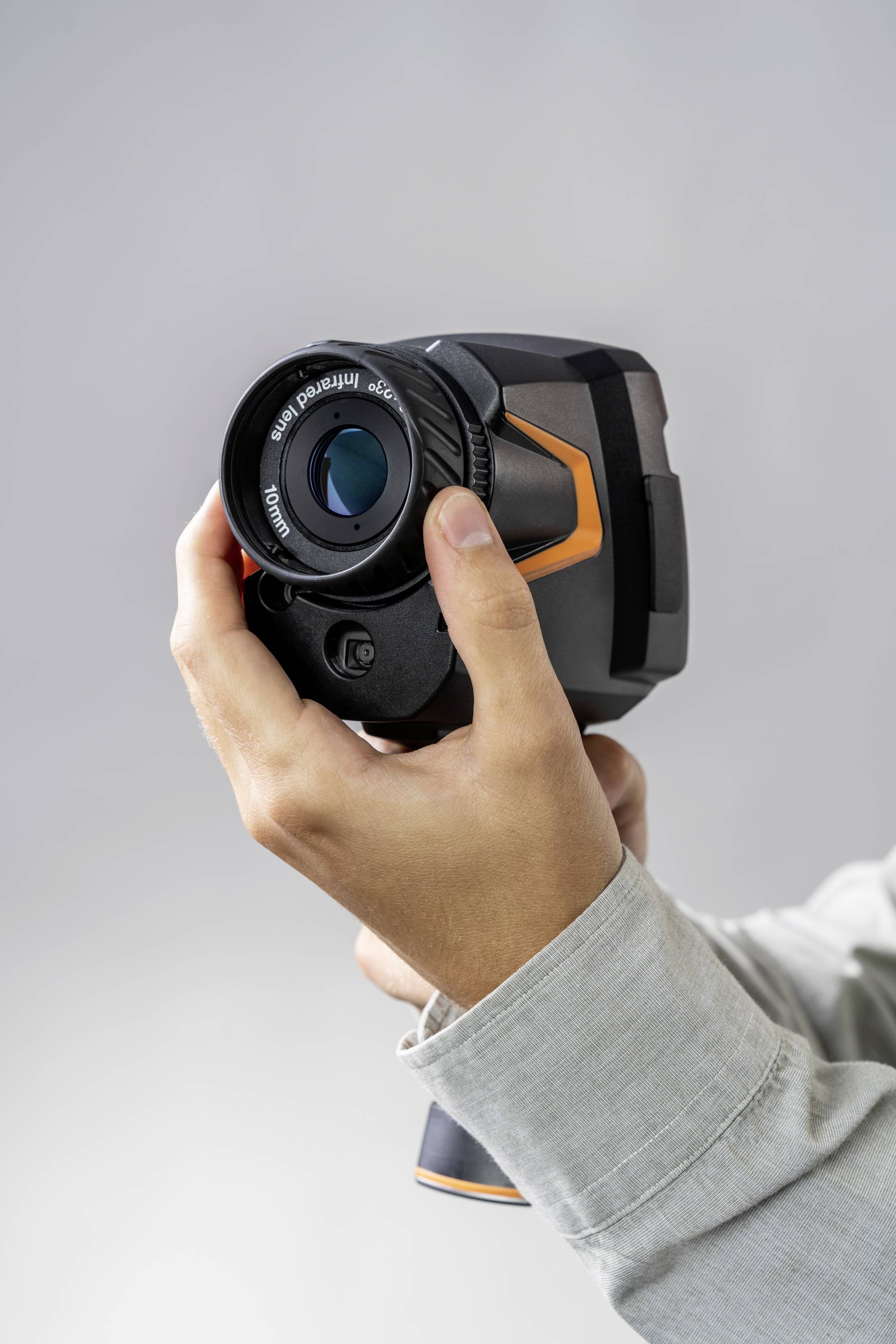 Una persona tiene una termocamera sullo sfondo grigio. La fotocamera ha un grande obiettivo e dettagli in arancione.