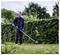 Una persona che indossa guanti e abbigliamento protettivo taglia una siepe utilizzando un tagliasiepi elettrico in un giardino con una vegetazione fitta.