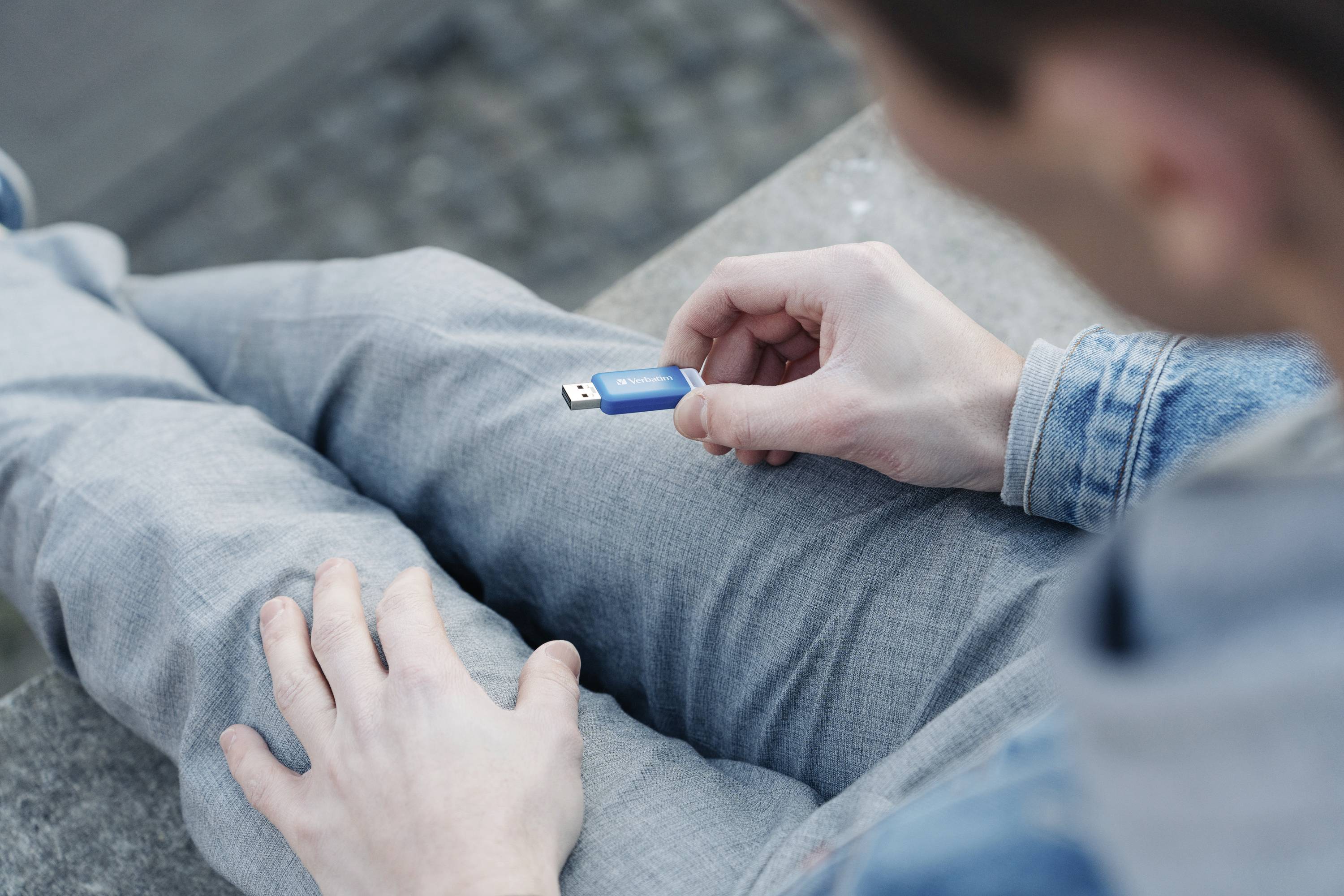 Una persona tiene un'unità flash USB blu in mano, seduta su un pantalone grigio. La scena è ripresa dall'alto.