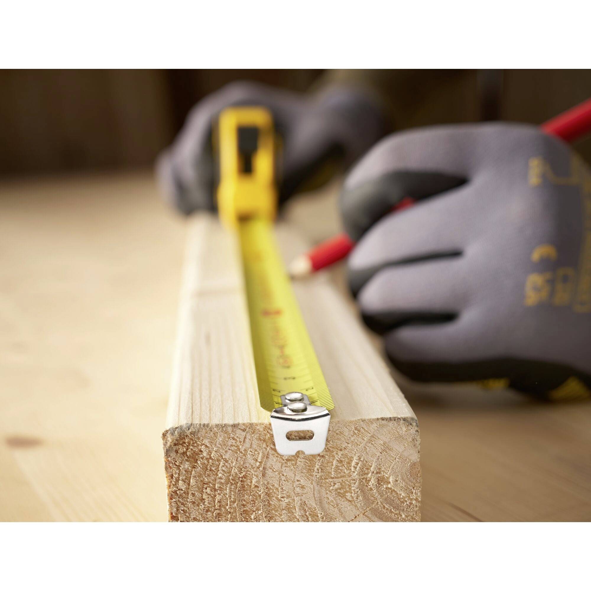 Una persona misura la lunghezza di un pezzo di legno con un nastro metrico giallo e una matita rossa. I guanti proteggono le mani.
