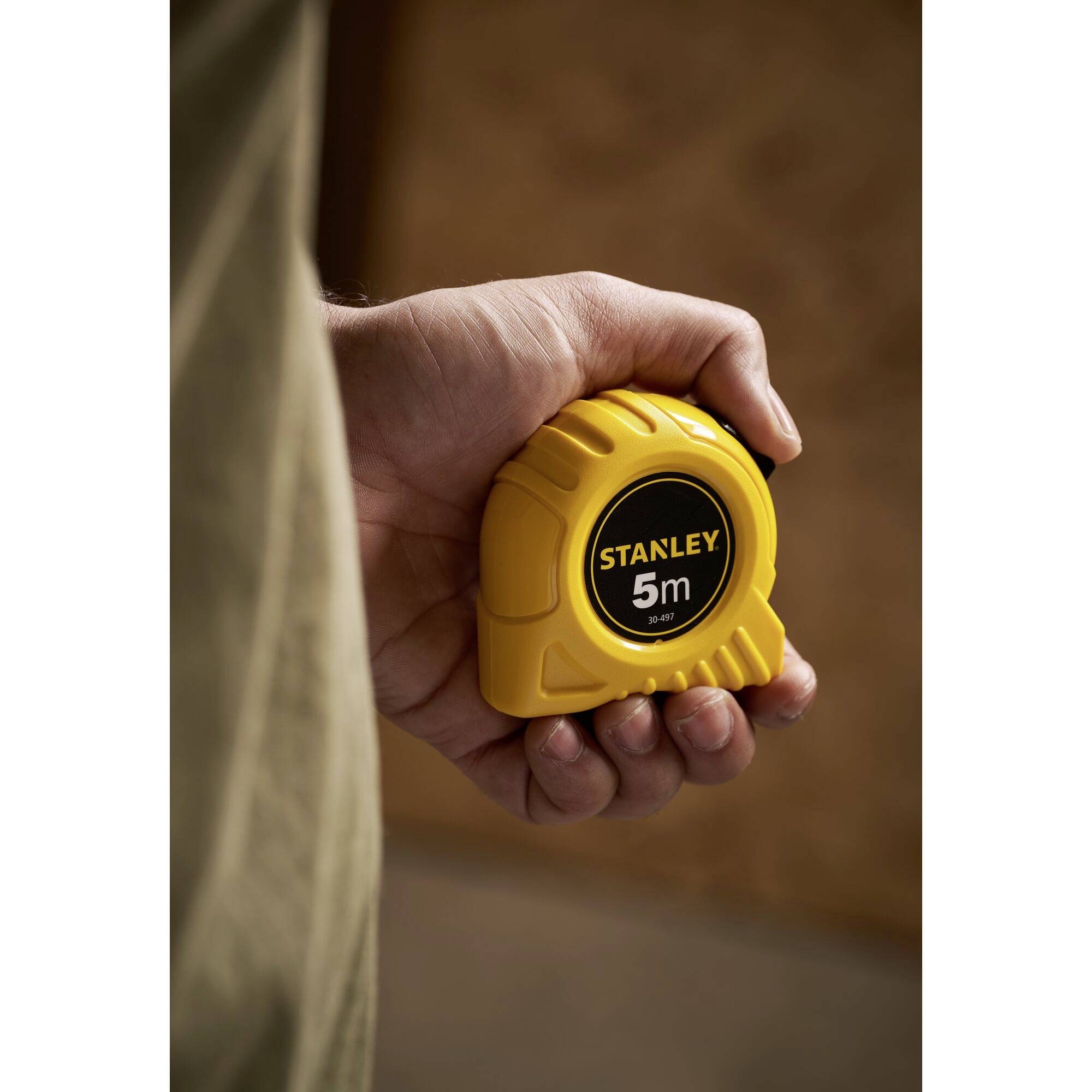 Una mano tiene un nastro metrico giallo Stanley con la scritta '5m', su uno sfondo sfocato, pronto per misurare.