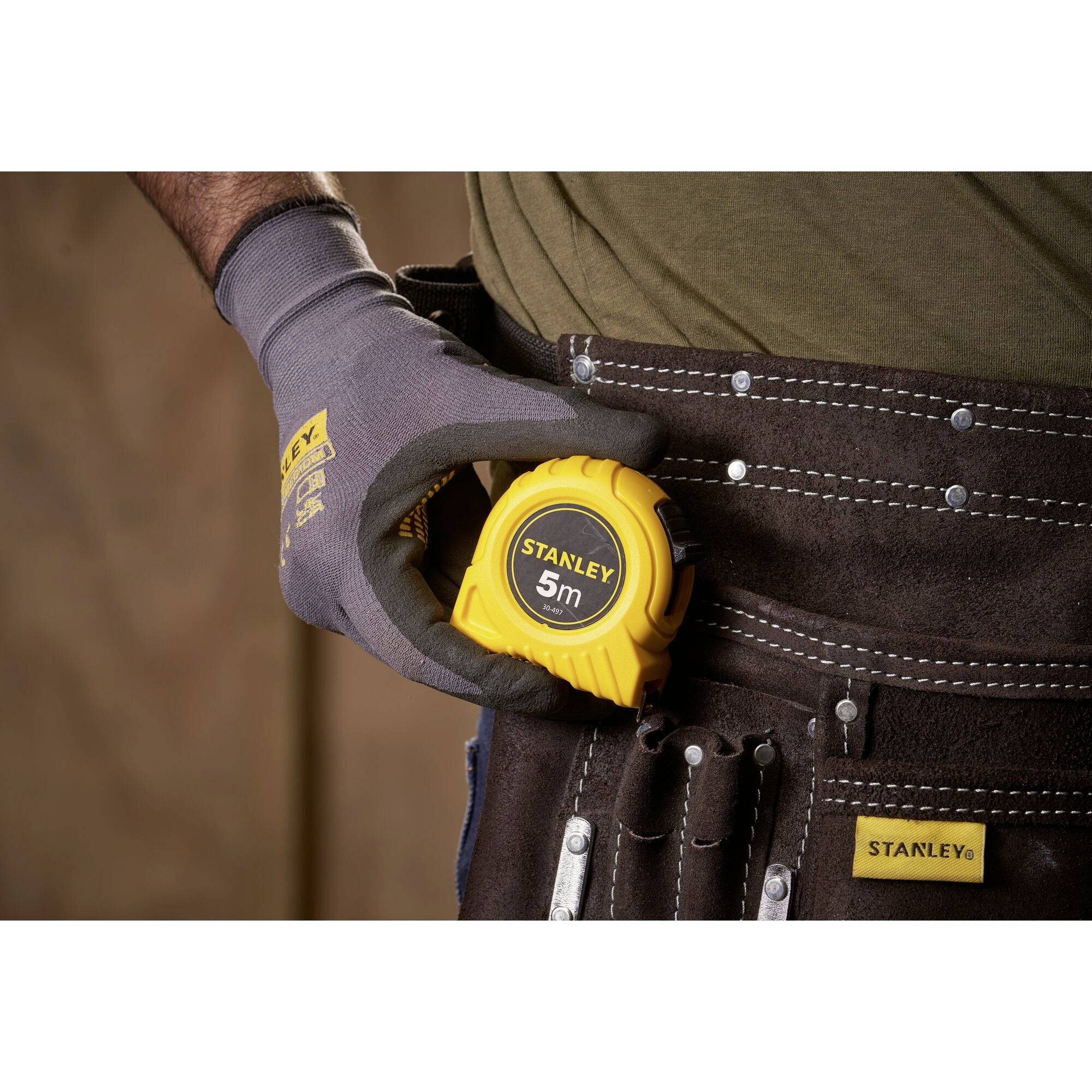 Una persona con abiti da lavoro tiene un nastro metrico giallo con la scritta 'STANLEY 5m' in una borsa degli attrezzi marrone.