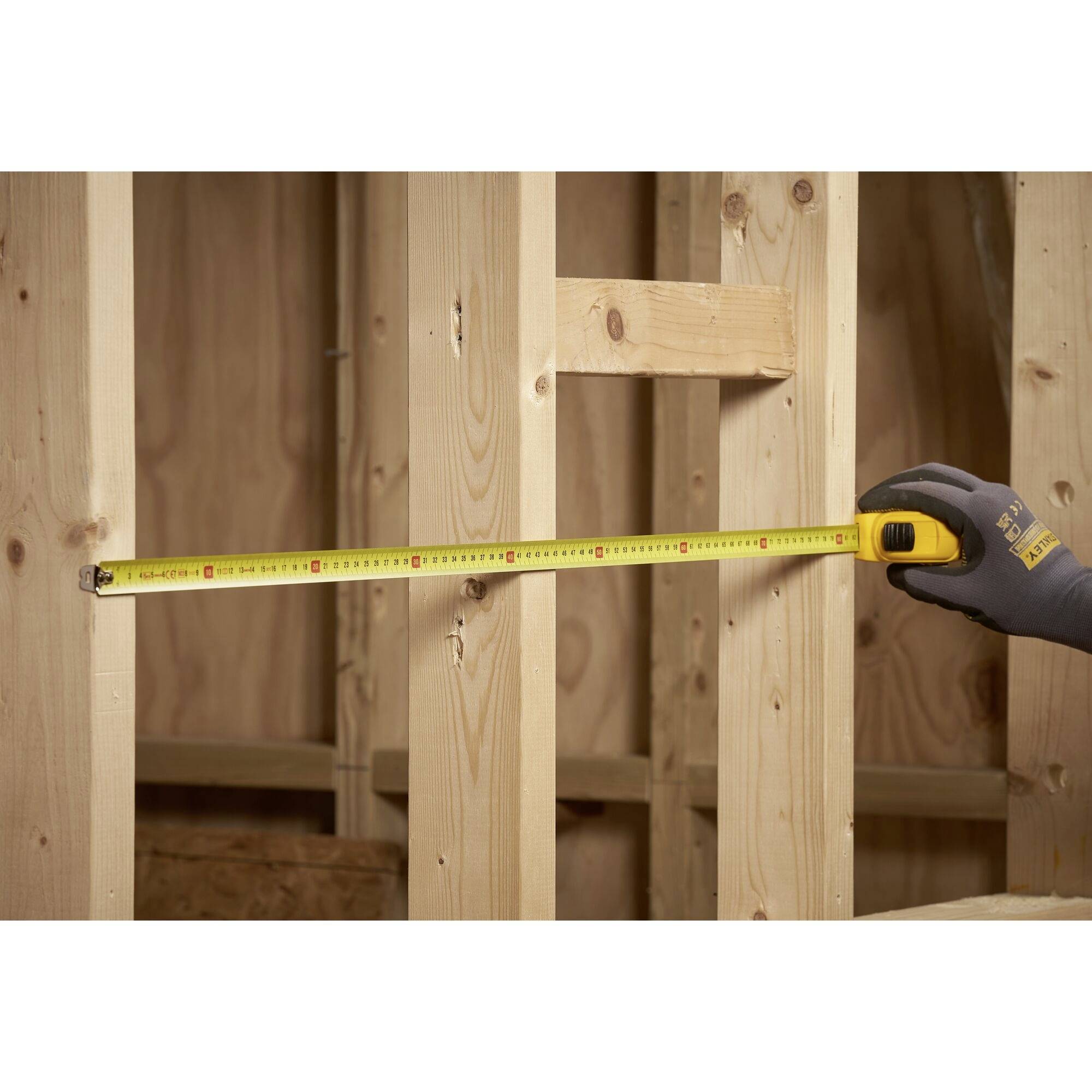 Una persona misura la lunghezza tra due pali di legno in un edificio in costruzione utilizzando un nastro metrico giallo.