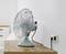 Un ventilatore da tavolo con custodia blu è posizionato su un tavolo di legno. È collegato a una presa a muro. Sullo sfondo si intravede un letto bianco.