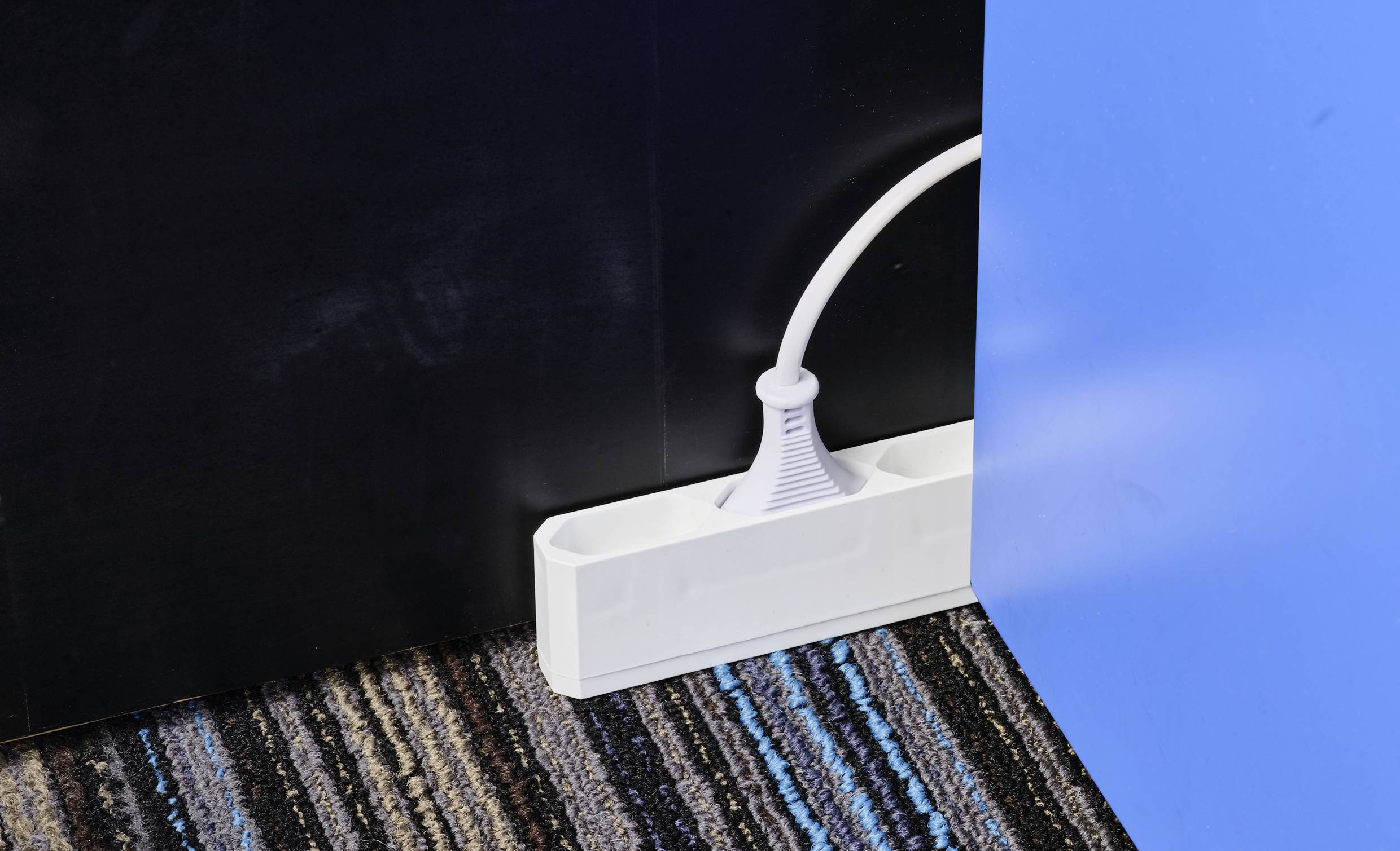 Un sistema di gestione dei cavi bianco è posizionato su un tappeto a righe e organizza un singolo cavo elettrico tra una parete nera e una blu.