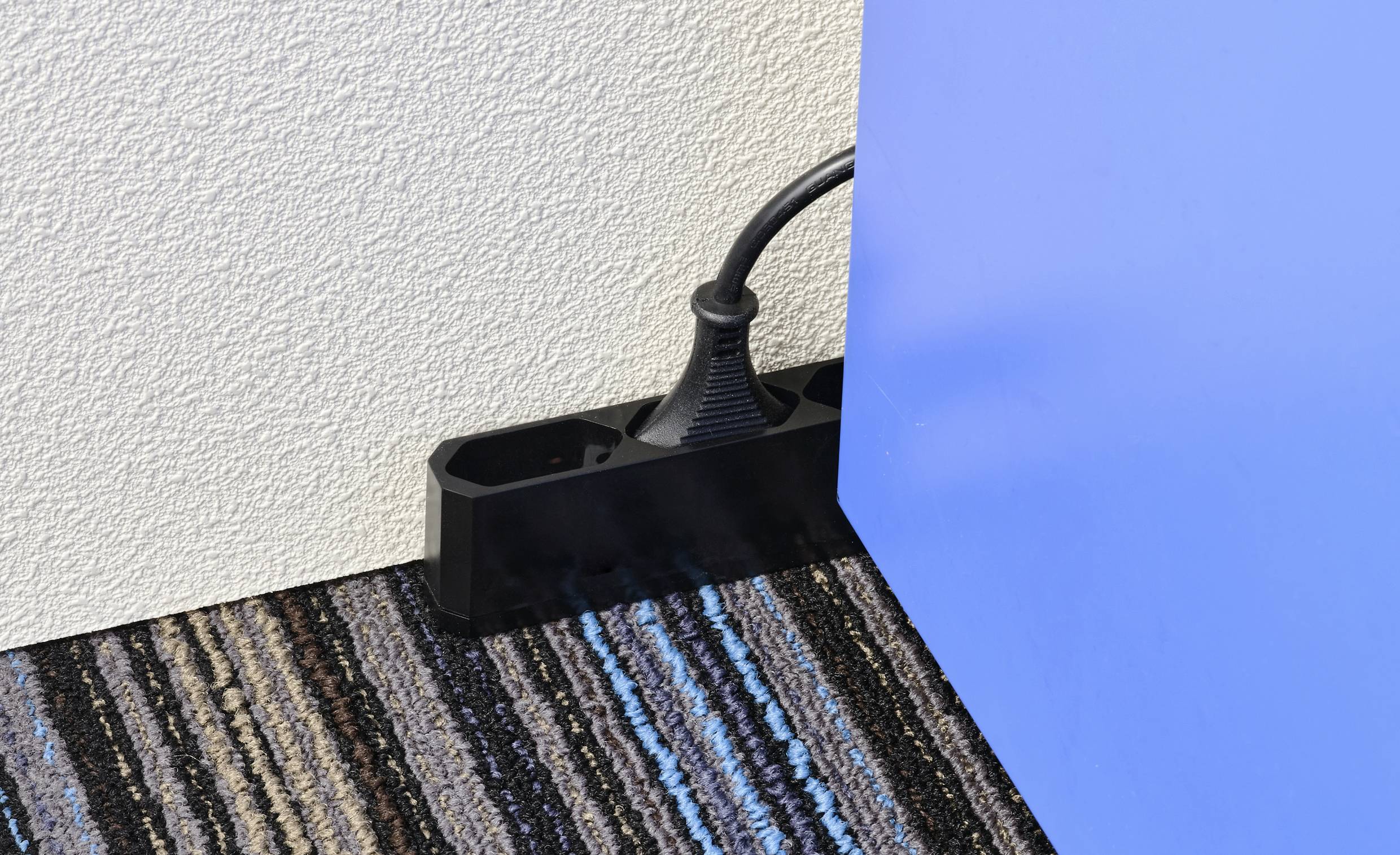 Multipresa nera posizionata dietro un tavolo blu, su un pavimento a righe, contro una parete bianca.