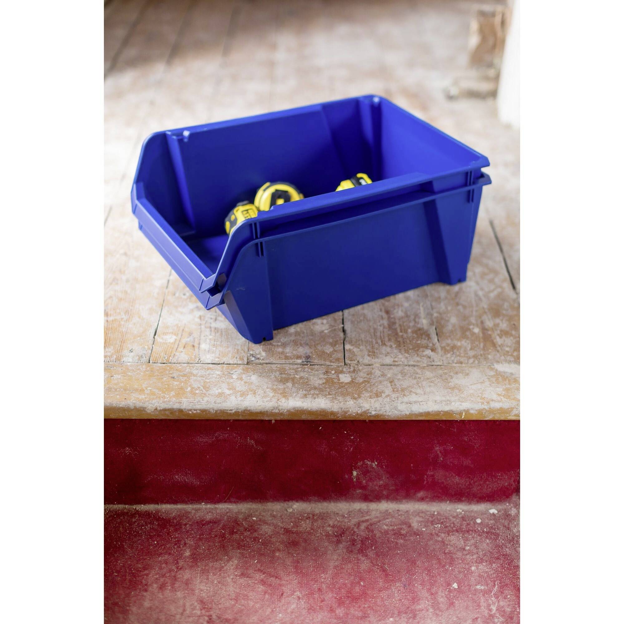 Una scatola di plastica blu con attrezzi gialli al suo interno è posizionata su un pavimento di legno consumato davanti a un gradino rosso.