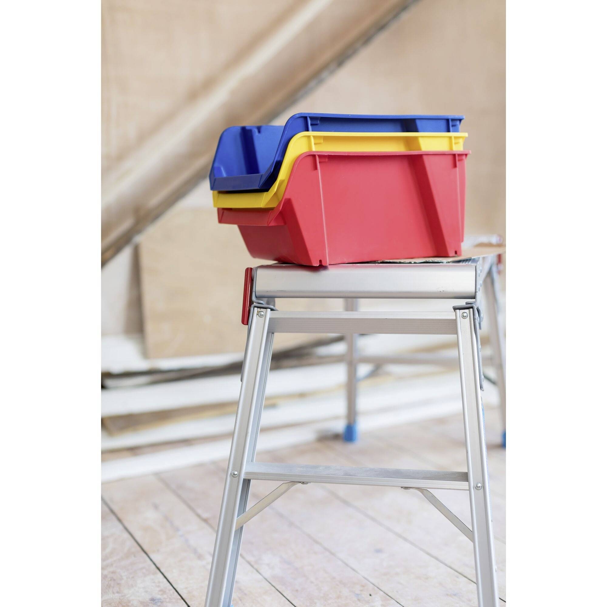 Diversi contenitori di plastica colorati sono impilati e posizionati su un piccolo tavolo, in una stanza con pavimento in legno e parete inclinata.